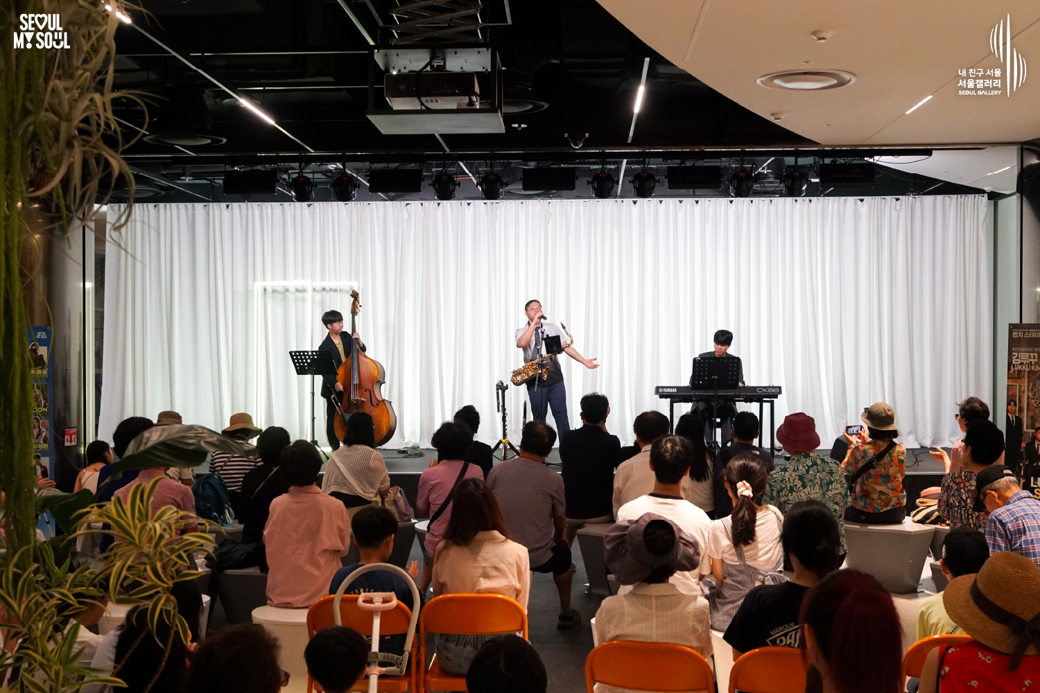 SEOUL MY SOUL 로고와 내 친구 서울 서울갤러리 SEOUL GALLERY 로고가 표기된 서울갤러리 공연장 사진으로, 흰색 커튼 배경의 무대 위에서 김루꾸재즈밴드의 세 남성 연주자가 콘트라베이스와 색소폰, 키보드를 연주하고 있으며, 객석에는 다양한 연령대의 관람객들이 앉아 공연을 관람하고 있는 모습이 담긴 이미지입니다.
