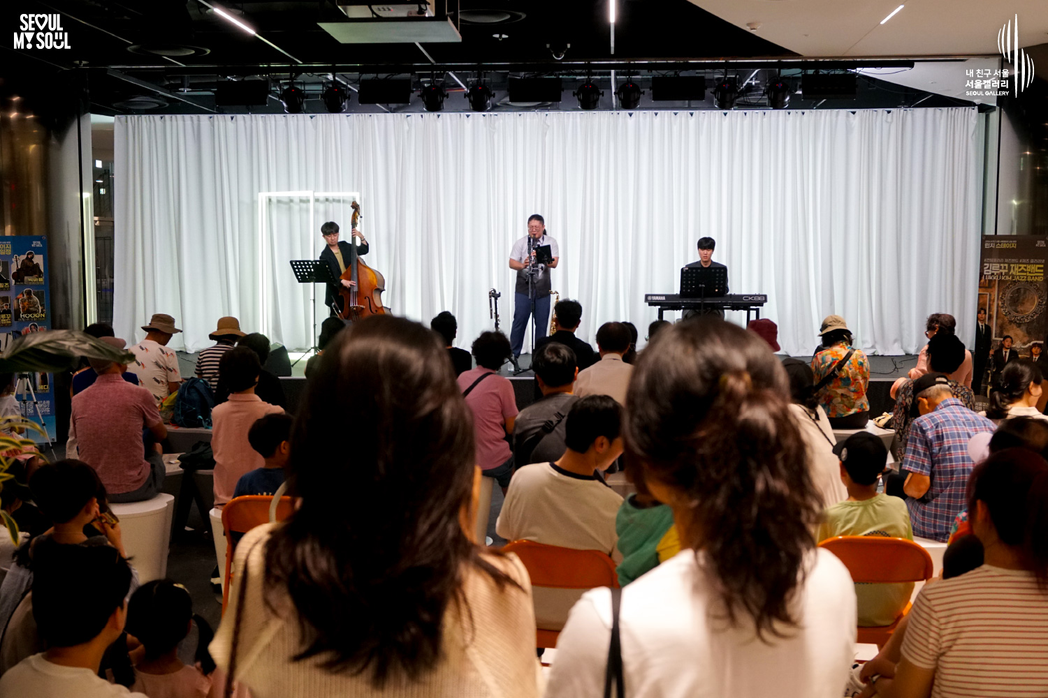SEOUL MY SOUL 로고와 내 친구 서울 서울갤러리 SEOUL GALLERY 로고가 표기된 서울갤러리 공연장 사진으로, 천장 조명이 설치된 흰색 커튼 배경의 무대에서 김루꾸재즈밴드가 콘트라베이스와 관악기, 키보드로 공연하고 있으며, 전경에는 여러 관람객의 뒷모습과 객석을 가득 채운 관람객들이 보이는 이미지입니다.