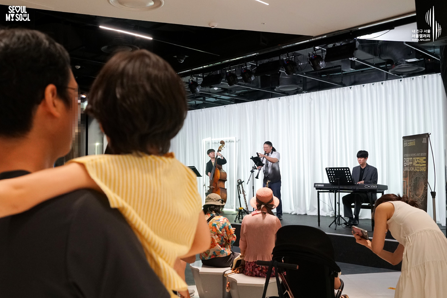 SEOUL MY SOUL 로고와 내 친구 서울 서울갤러리 SEOUL GALLERY 로고가 표기된 서울갤러리 공연장 사진으로, 흰색 커튼 배경의 무대에서 세 남성 연주자가 콘트라베이스와 관악기, 키보드로 공연하고 있으며, 왼쪽 전경에는 아이를 안고 공연을 바라보는 관람객이 크게 보이고 객석에도 여러 관람객이 앉아 있는 이미지입니다.