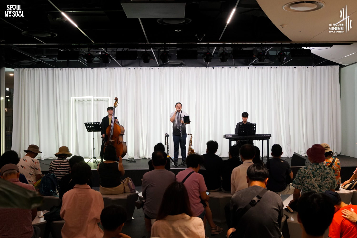 SEOUL MY SOUL 로고와 내 친구 서울 서울갤러리 SEOUL GALLERY 로고가 표기된 서울갤러리 공연장 사진으로, 천장 무대 조명 아래 흰색 커튼 배경의 무대에서 김루꾸재즈밴드의 남성 연주자들이 콘트라베이스와 관악기, 키보드로 공연하고 있으며, 객석에는 다양한 연령대의 관람객들이 앉아 무대를 바라보고 있는 모습이 담긴 이미지입니다.
