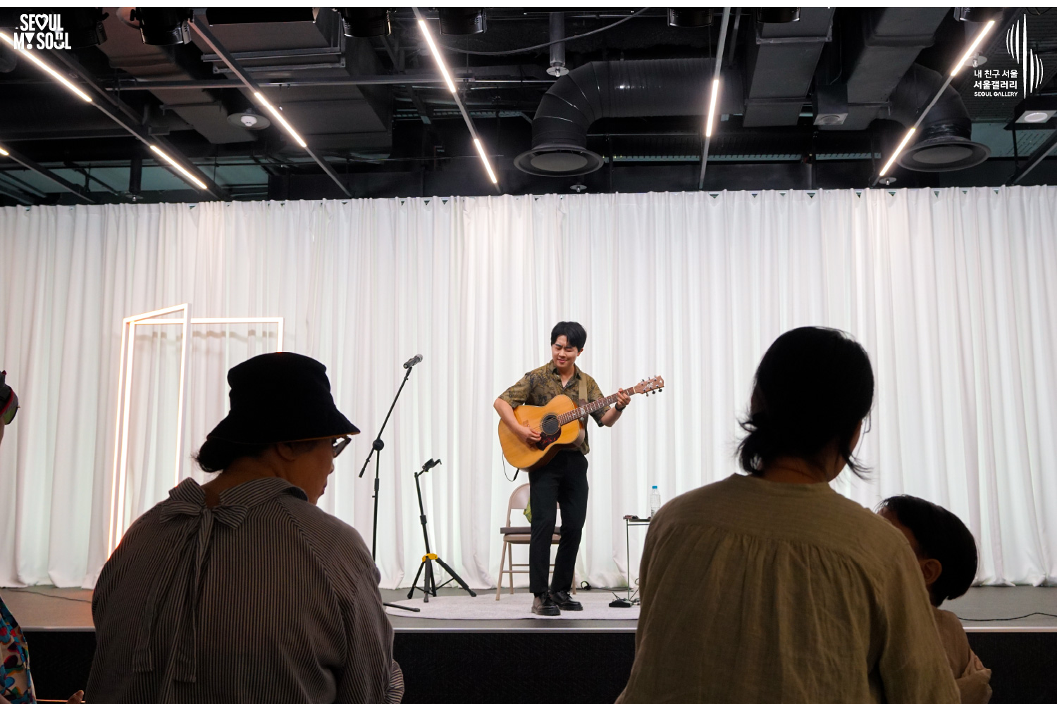SEOUL MY SOUL 로고와 내 친구 서울 서울갤러리 SEOUL GALLERY 로고가 표기된 서울갤러리 공연장 사진으로, 흰색 커튼과 사각 조명이 설치된 무대 위에서 셔츠와 바지를 입은 남성 기타리스트가 어쿠스틱 기타를 연주하고 있으며, 전경에는 두 관람객의 뒷모습이 크게 보이는 모습이 담긴 이미지입니다.