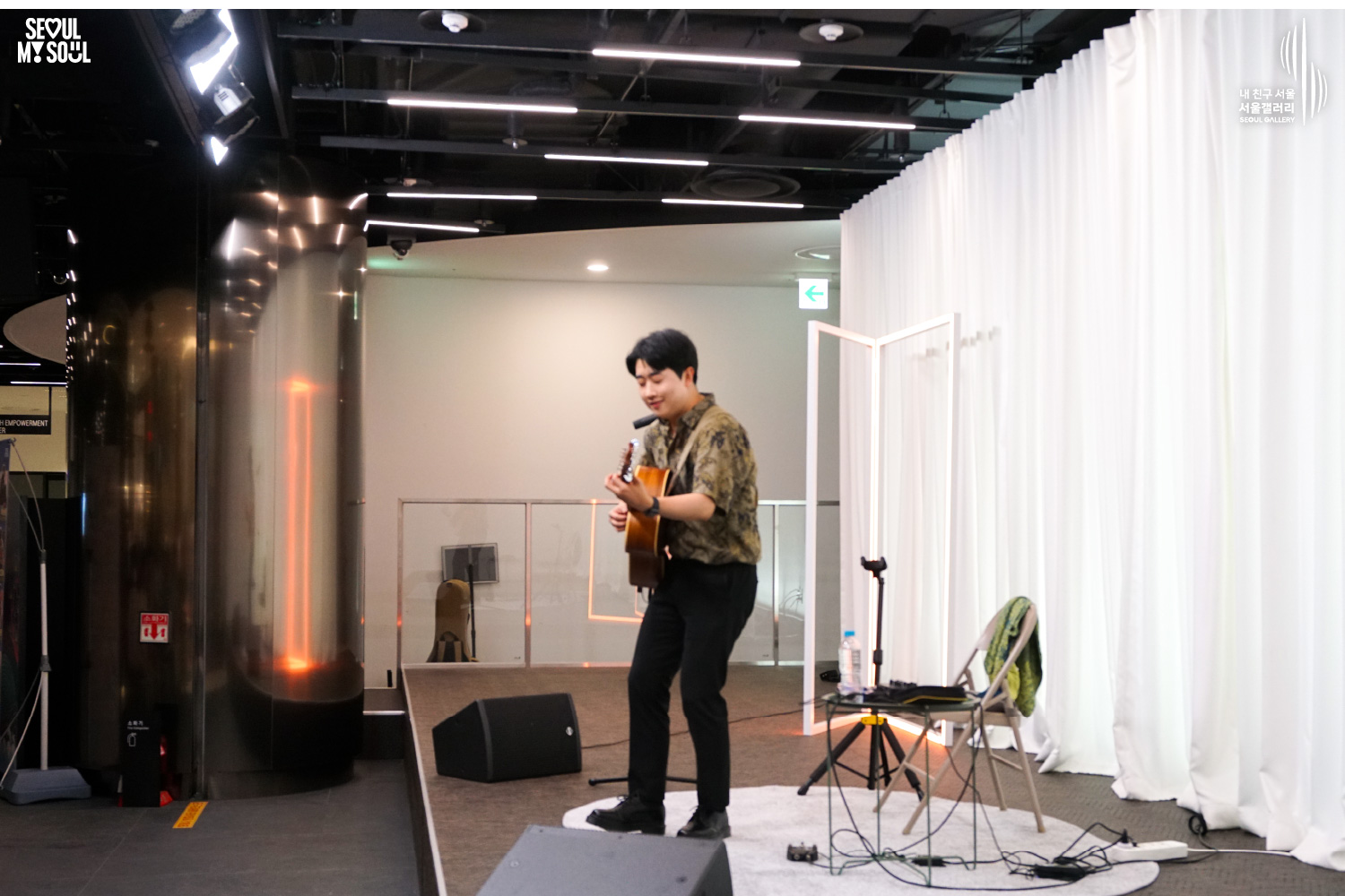 SEOUL MY SOUL 로고와 내 친구 서울 서울갤러리 SEOUL GALLERY 로고가 표기된 서울갤러리 공연장 무대 사진으로, 흰색 커튼과 사각 조명이 있는 무대 위에서 셔츠와 바지를 입은 남성 기타리스트가 어쿠스틱 기타를 연주하고 있으며, 주변에는 마이크 스탠드와 의자, 작은 테이블, 모니터 스피커가 놓여 있는 이미지입니다.