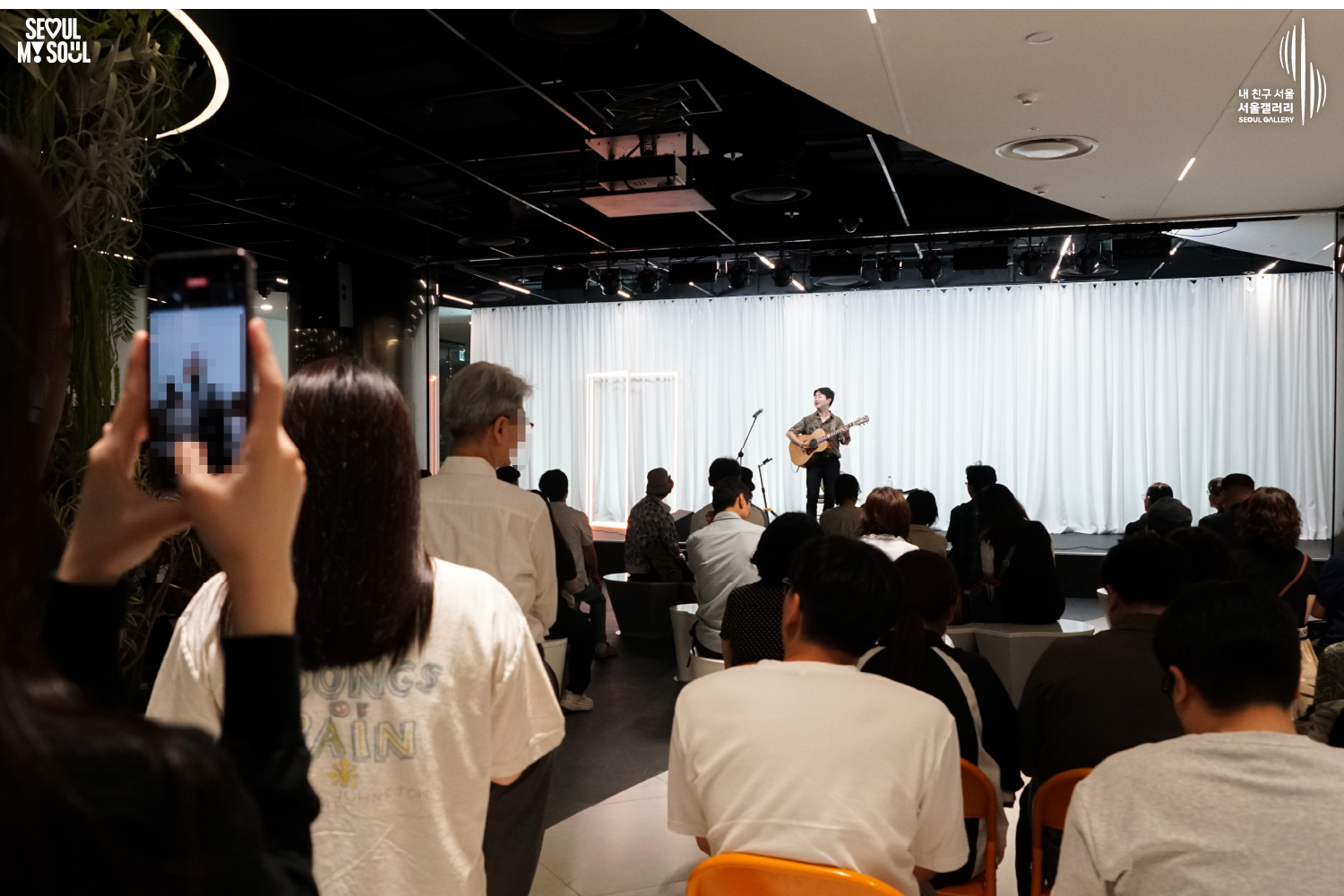 SEOUL MY SOUL 로고와 내 친구 서울 서울갤러리 SEOUL GALLERY 로고가 표기된 서울갤러리 지하 1층 공연장의 공연 현장 사진으로, 흰색 커튼 배경의 무대에서 남성 기타리스트가 어쿠스틱 기타를 연주하고 있으며, 객석에는 여러 관람객이 앉아 있고 왼쪽 전경에서는 한 관람객이 휴대전화로 무대를 촬영하고 있는 모습이 담긴 이미지입니다.