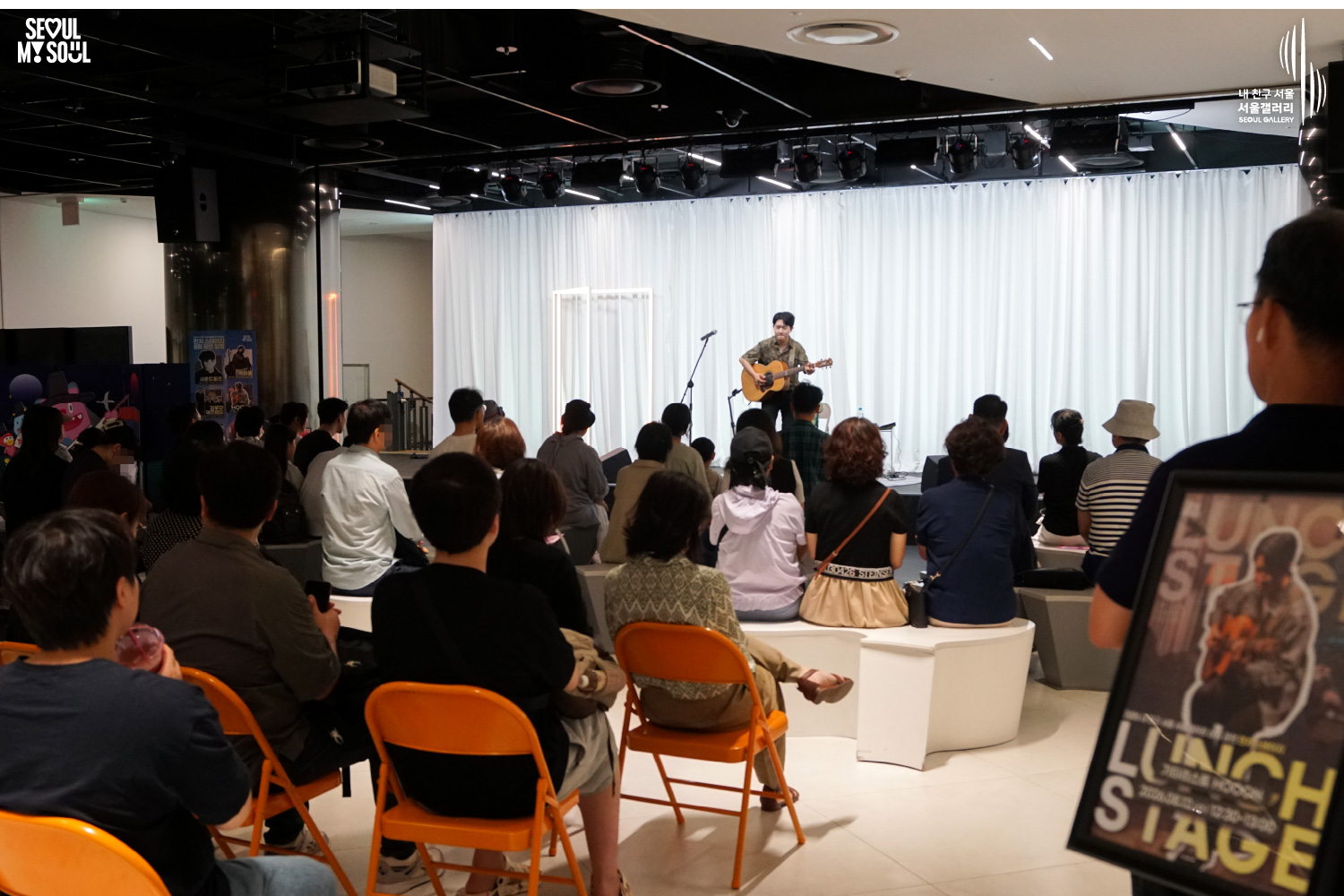 SEOUL MY SOUL 로고와 내 친구 서울 서울갤러리 SEOUL GALLERY 로고가 표기된 서울갤러리 지하 1층 공연장의 공연 현장 사진으로, 천장 조명 아래 흰색 커튼 배경의 무대에서 남성 기타리스트가 어쿠스틱 기타를 연주하고 있으며, 객석에는 많은 관람객이 앉아 있고 오른쪽 전경에는 공연 안내판이 보이는 이미지입니다.