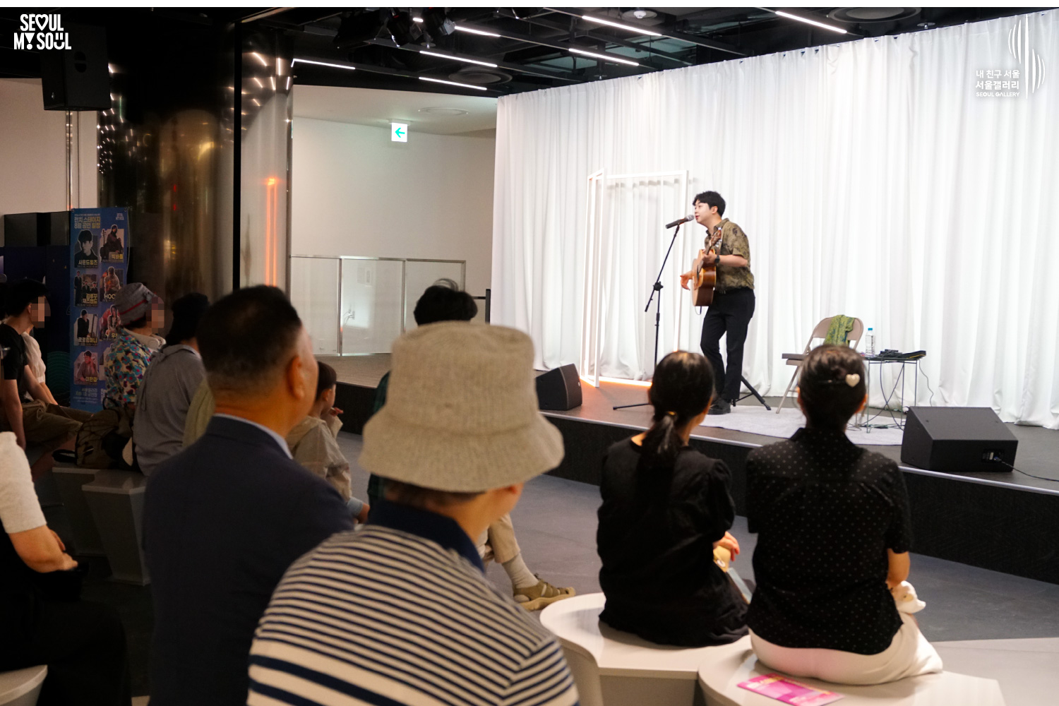 SEOUL MY SOUL 로고와 내 친구 서울 서울갤러리 SEOUL GALLERY 로고가 표기된 서울갤러리 공연장 사진으로, 흰색 커튼과 사각 조명이 설치된 무대 위에서 셔츠와 바지를 입은 남성 기타리스트가 어쿠스틱 기타를 메고 마이크 앞에서 노래를 부르고 있으며, 앞쪽 객석의 관람객들이 무대를 바라보고 있는 모습이 담긴 이미지입니다.