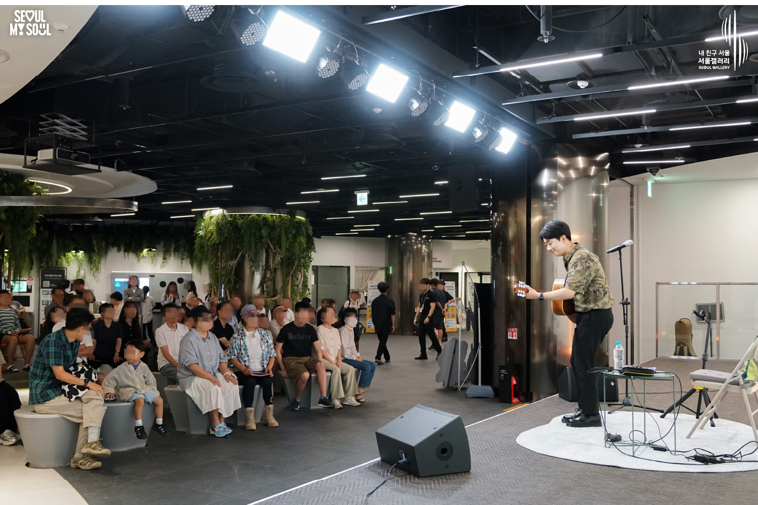 SEOUL MY SOUL 로고와 내 친구 서울 서울갤러리 SEOUL GALLERY 로고가 표기된 서울갤러리 지하 1층 공연장의 공연 현장 사진으로, 천장에 여러 조명이 설치된 무대에서 남성 기타리스트가 어쿠스틱 기타를 연주하고 있으며, 무대 옆과 정면 객석에는 어린이를 포함한 여러 관람객들이 앉아 공연을 관람하고 있는 모습이 담긴 이미지입니다.