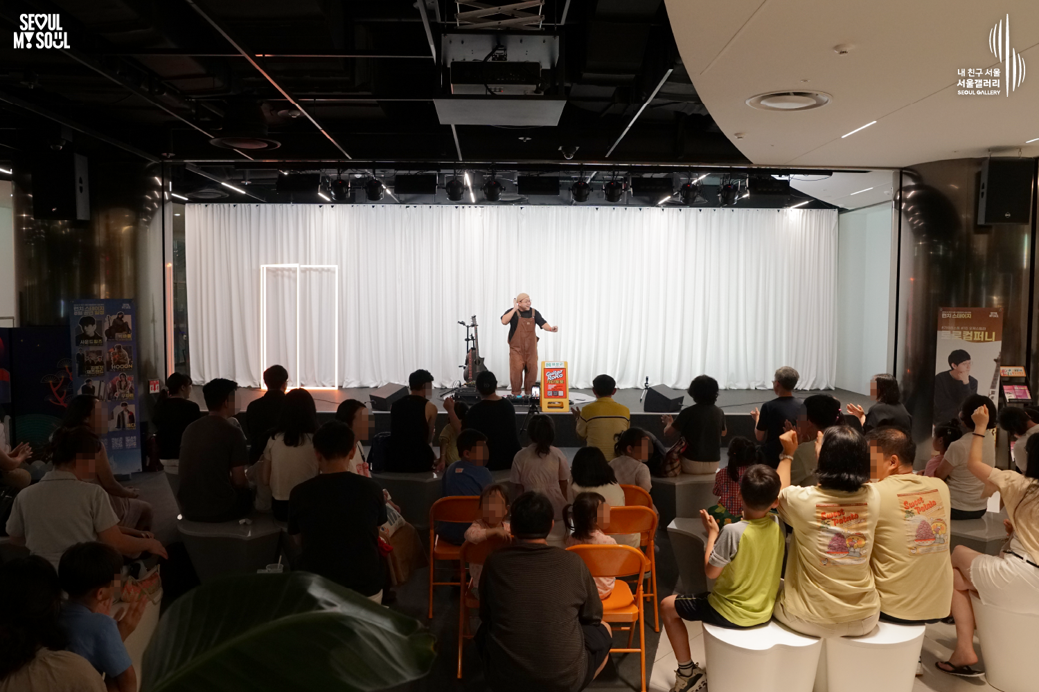 SEOUL MY SOUL 로고와 내 친구 서울 서울갤러리 SEOUL GALLERY 로고가 표기된 서울갤러리 지하 1층 공연장의 공연 현장 사진으로, 천장 무대 조명이 설치된 흰색 커튼 배경의 무대 위에서 모자와 안경을 착용하고 멜빵바지를 입은 아티스트가 한 손을 귀에 대고 다른 손을 앞으로 내밀고 있으며, 무대에는 악기와 스피커, 공연 안내판이 놓여 있고 객석에는 어린이를 포함한 다양한 연령대의 관람객들이 앉아 무대를 바라보는 모습이 담긴 이미지입니다.