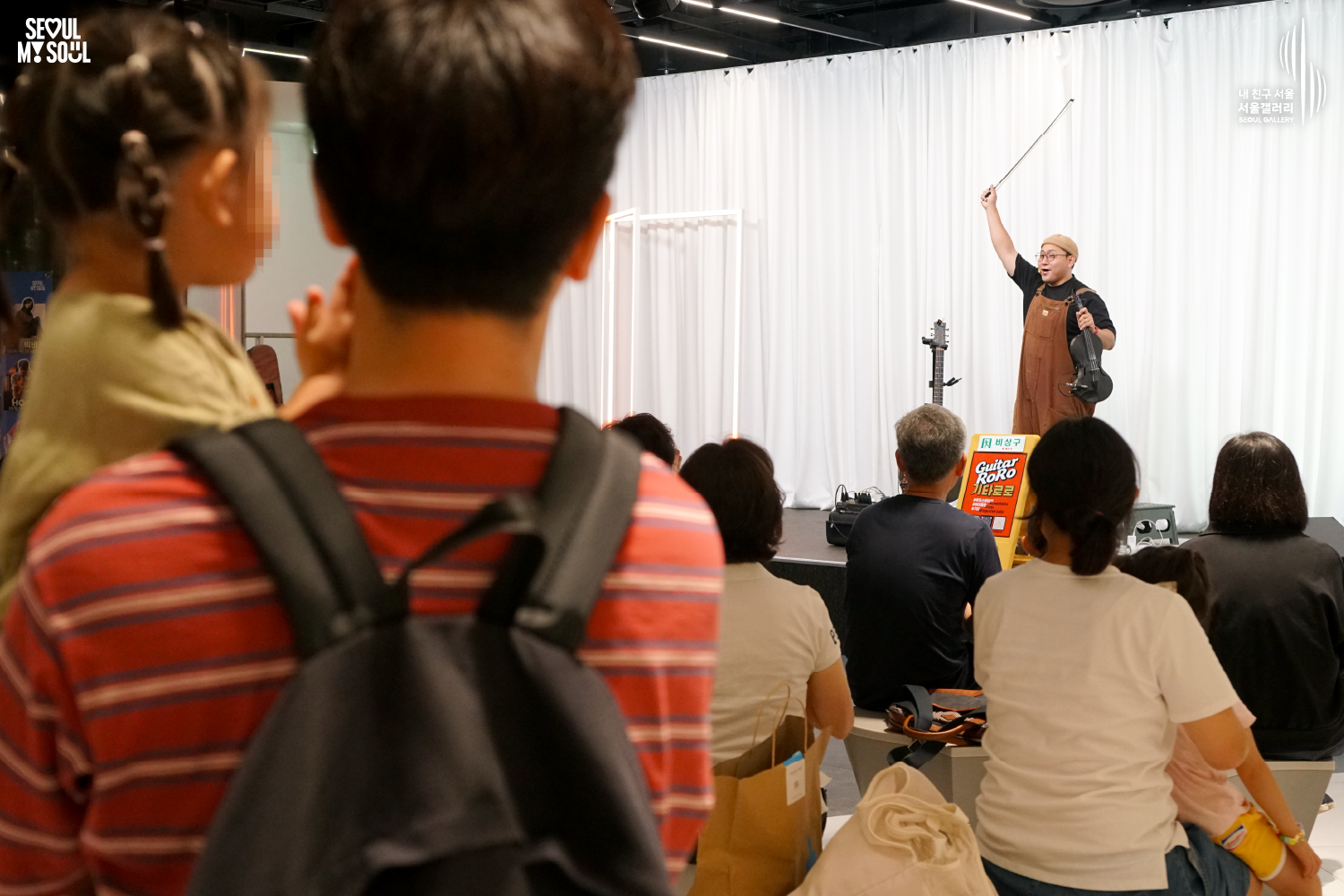 SEOUL MY SOUL 로고와 내 친구 서울 서울갤러리 SEOUL GALLERY 로고가 표기된 서울갤러리 공연장 사진으로, 천장 무대 조명이 비추는 흰색 커튼 배경의 무대 위에서 모자와 안경을 착용하고 멜빵바지를 입은 아티스트가 한 손에 바이올린을 들고 다른 손으로 활을 높이 들어 올리고 있으며, 공연 안내판과 악기가 놓인 무대 앞 객석에서 관람객들이 무대를 바라보는 모습이 담긴 이미지입니다.