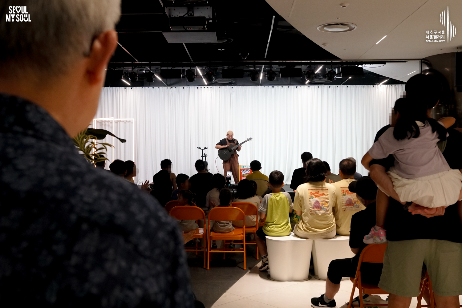 SEOUL MY SOUL 로고와 내 친구 서울 서울갤러리 SEOUL GALLERY 로고가 표기된 서울갤러리 공연장 사진으로, 천장 무대 조명이 설치된 흰색 커튼 배경의 무대 위에서 모자와 안경을 착용하고 멜빵바지를 입은 아티스트가 기타를 연주하고 있으며, 객석 뒤편에서 무대를 바라본 구도로 앞쪽에는 여러 관람객의 뒷모습과 어린아이를 안고 서 있는 관람객이 함께 보이는 모습이 담긴 이미지입니다.