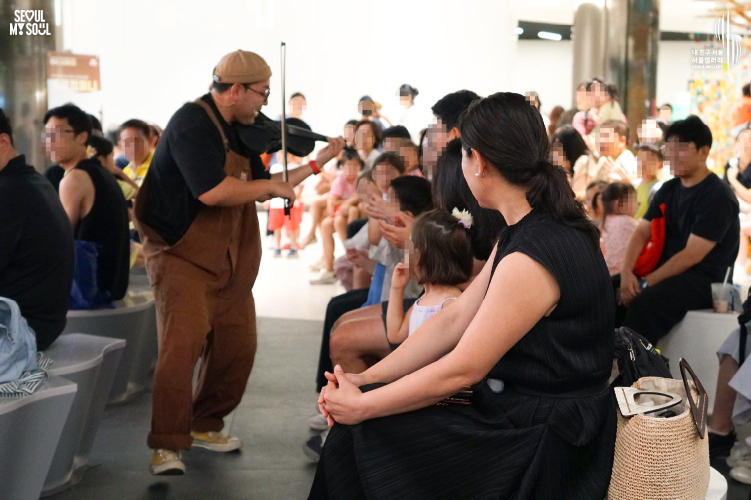 SEOUL MY SOUL 로고와 내 친구 서울 서울갤러리 SEOUL GALLERY 로고가 표기된 서울갤러리 공연장 사진으로, 천장 조명이 밝게 비추는 실내 공간에서 모자와 안경을 착용하고 멜빵바지를 입은 아티스트가 객석 사이에 서서 바이올린을 활로 연주하고 있으며, 가까이 앉아 있는 어린이와 성인 관람객들이 연주자를 바라보며 공연을 관람하는 모습이 담긴 이미지입니다.