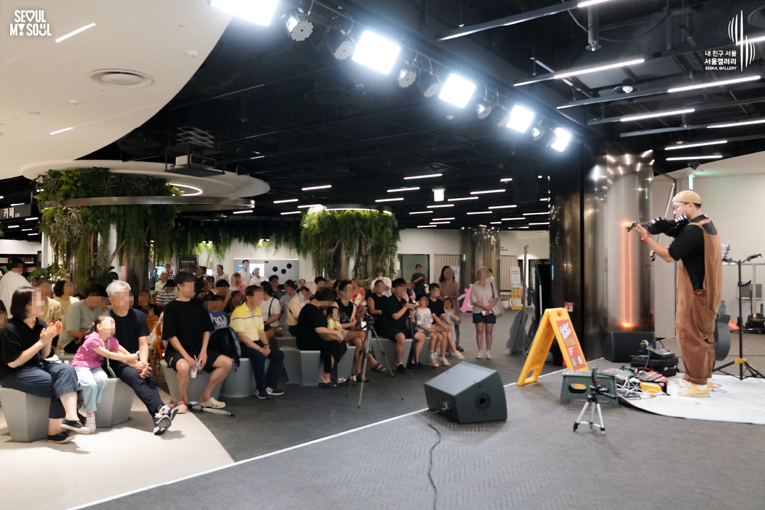 SEOUL MY SOUL 로고와 내 친구 서울 서울갤러리 SEOUL GALLERY 로고가 표기된 서울갤러리 지하 1층 공연장의 공연 현장 사진으로, 천장의 밝은 무대 조명이 비추는 실내 공간에서 모자와 안경을 착용하고 멜빵바지를 입은 아티스트가 무대 한쪽에서 바이올린을 활로 연주하고 있으며, 무대에는 스피커와 악기 장비가 놓여 있고 객석에는 어린이와 성인 관람객들이 앉거나 서서 공연을 관람하는 모습이 담긴 이미지입니다.