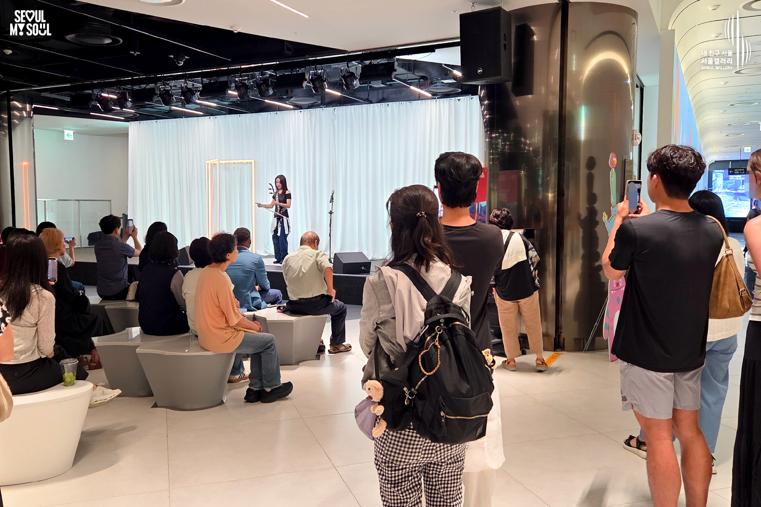 SEOUL MY SOUL 로고와 내 친구 서울 서울갤러리 SEOUL GALLERY 로고가 표기된 서울갤러리 지하 1층 공연장의 공연 현장 사진으로, 천장 무대 조명이 비추는 흰색 커튼 배경의 무대 위에서 긴 머리의 여성 연주자가 해금을 들고 공연하고 있으며, 객석에는 관람객들이 앉아 있고 통로와 무대 주변에는 서서 공연을 바라보거나 휴대전화로 촬영하는 관람객들이 함께 보이는 모습이 담긴 이미지입니다.