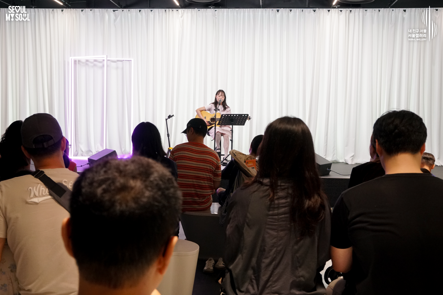 SEOUL MY SOUL 로고와 내 친구 서울 서울갤러리 SEOUL GALLERY 로고가 표기된 서울갤러리 공연장 사진으로, 보랏빛 조명과 사각 조명이 어우러진 흰색 커튼 배경의 무대 위에서 긴 머리에 원피스를 입은 여성 싱어송라이터가 의자에 앉아 어쿠스틱 기타를 연주하며 마이크 앞에서 노래하고 있으며, 앞에는 악보대와 모니터 스피커가 놓여 있고 객석에는 관람객들이 무대를 바라보는 모습이 담긴 이미지입니다.
