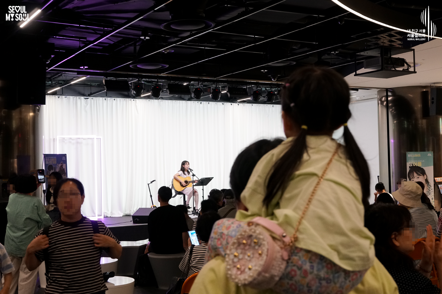 SEOUL MY SOUL 로고와 내 친구 서울 서울갤러리 SEOUL GALLERY 로고가 표기된 서울갤러리 공연장 사진으로, 천장 무대 조명과 보랏빛 조명이 비추는 흰색 커튼 배경의 무대에서 긴 머리에 원피스를 입은 여성 싱어송라이터가 의자에 앉아 어쿠스틱 기타를 연주하며 마이크 앞에서 노래하고 있으며, 객석에는 많은 관람객들이 공연을 관람하고 전경에는 어린아이를 안고 무대를 바라보는 관람객이 크게 보이는 모습이 담긴 이미지입니다.