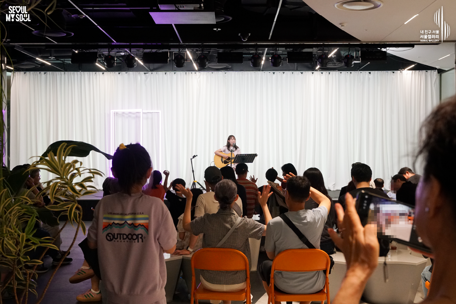 SEOUL MY SOUL 로고와 내 친구 서울 서울갤러리 SEOUL GALLERY 로고가 표기된 서울갤러리 공연장 사진으로, 천장 무대 조명과 보랏빛 조명이 비추는 흰색 커튼 배경의 무대에서 긴 머리에 원피스를 입은 여성 싱어송라이터가 어쿠스틱 기타를 연주하며 마이크 앞에서 노래하고 있으며, 객석에는 다양한 연령대의 관람객들이 앉거나 서서 손을 들어 공연에 참여하고 일부는 휴대전화로 무대를 촬영하는 모습이 담긴 이미지입니다.