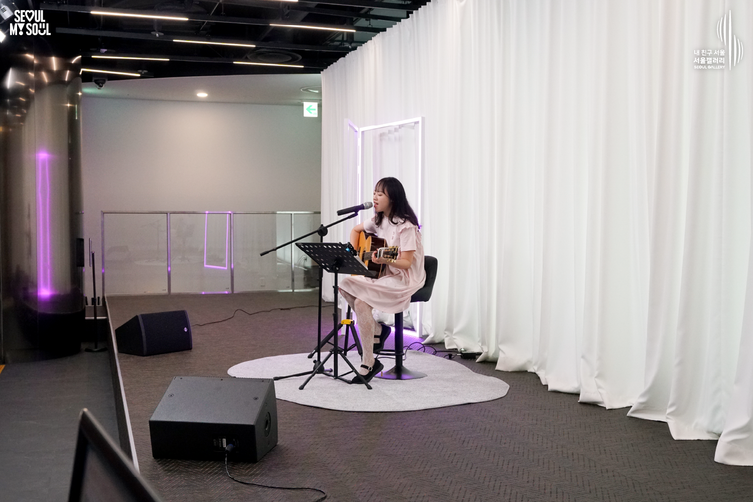 SEOUL MY SOUL 로고와 내 친구 서울 서울갤러리 SEOUL GALLERY 로고가 표기된 서울갤러리 공연장 무대 사진으로, 보랏빛 조명과 사각 조명이 비추는 흰색 커튼 배경의 무대 위에서 긴 머리에 원피스를 입은 여성 싱어송라이터가 의자에 앉아 어쿠스틱 기타를 연주하며 마이크 앞에서 노래하고 있으며, 앞에는 악보대와 마이크 스탠드, 모니터 스피커가 놓여 있는 모습이 담긴 이미지입니다.