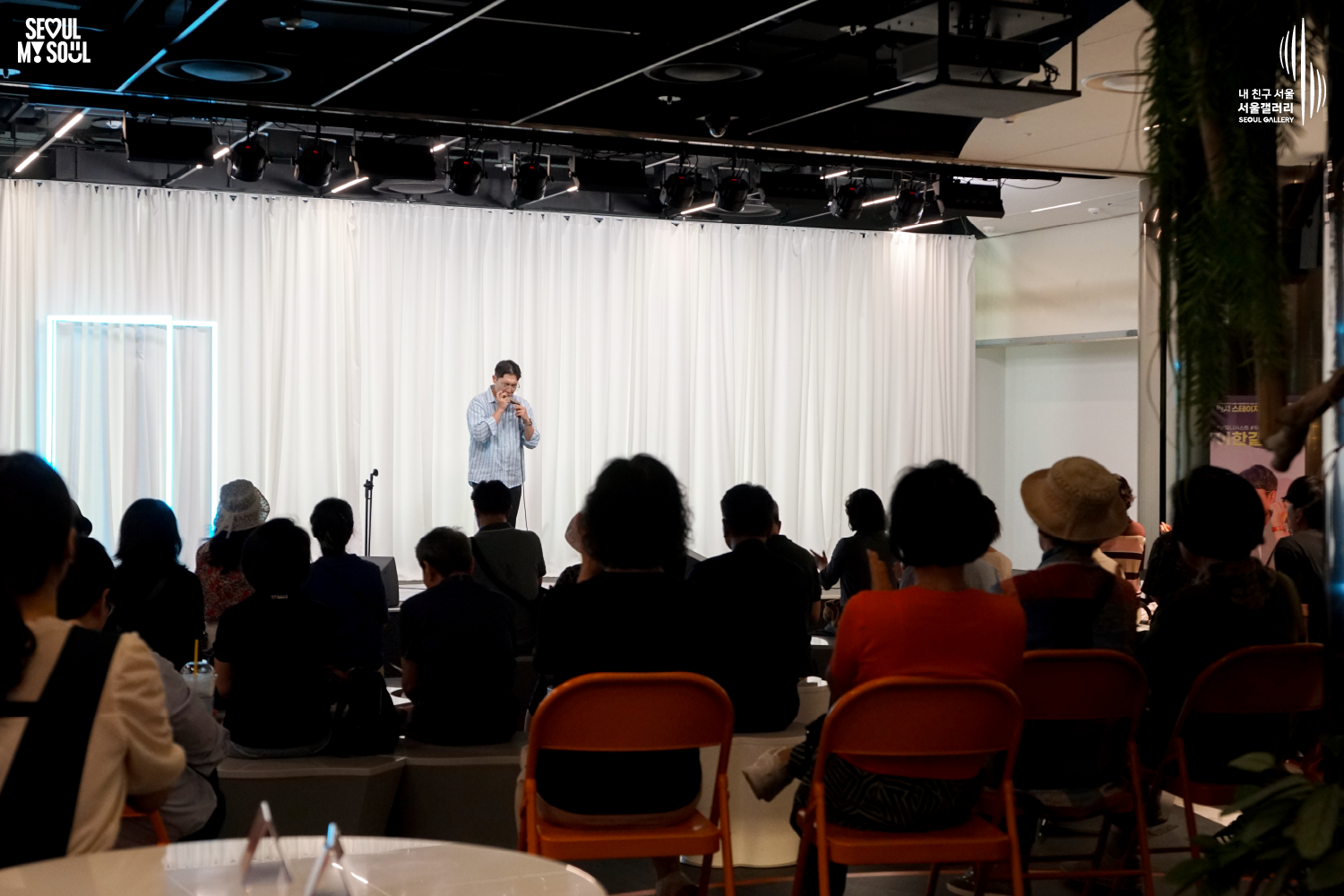 SEOUL MY SOUL 로고와 내 친구 서울 서울갤러리 SEOUL GALLERY 로고가 표기된 서울갤러리 공연장 사진으로, 천장 무대 조명과 푸른빛 사각 조명이 비추는 흰색 커튼 배경의 무대 위에서 짧은 머리에 안경을 착용하고 셔츠와 바지를 입은 남성 연주자가 하모니카를 두 손으로 감싸 입에 대고 연주하고 있으며, 객석에는 다양한 연령대의 관람객들이 앉아 무대를 바라보는 모습이 담긴 이미지입니다.