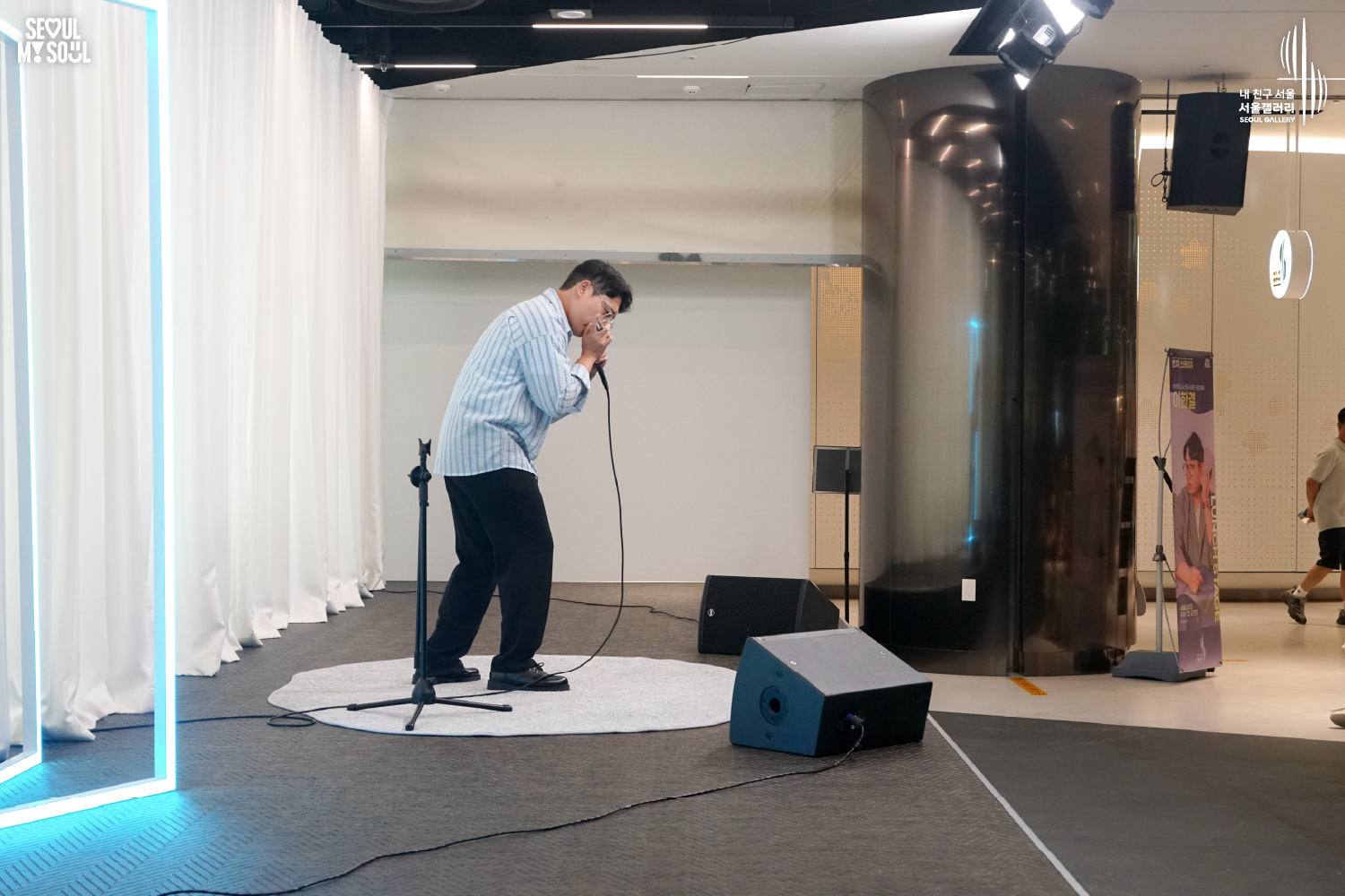 SEOUL MY SOUL 로고와 내 친구 서울 서울갤러리 SEOUL GALLERY 로고가 표기된 서울갤러리 공연장 무대 사진으로, 푸른빛 사각 조명과 천장 조명이 비추는 흰색 커튼 배경의 무대 위에서 짧은 머리에 안경을 착용하고 셔츠와 바지를 입은 남성 연주자가 몸을 앞으로 숙인 채 하모니카를 입에 대고 연주하고 있으며, 무대에는 마이크 스탠드와 모니터 스피커가 놓여 있는 모습이 담긴 이미지입니다.