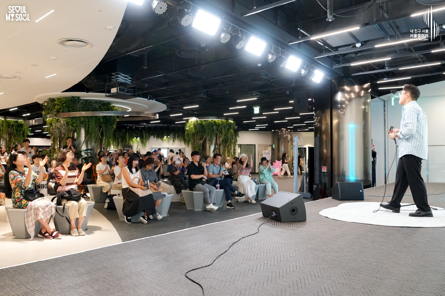 SEOUL MY SOUL 로고와 내 친구 서울 서울갤러리 SEOUL GALLERY 로고가 표기된 서울갤러리 지하 1층 공연장의 공연 현장 사진으로, 천장의 밝은 무대 조명과 푸른빛 조명이 비추는 실내 공간에서 짧은 머리에 안경을 착용하고 셔츠와 바지를 입은 남성 연주자가 한 손에 마이크를 들고 객석 방향을 바라보고 있으며, 객석의 관람객들은 손뼉을 치며 무대를 바라보는 모습이 담긴 이미지입니다.