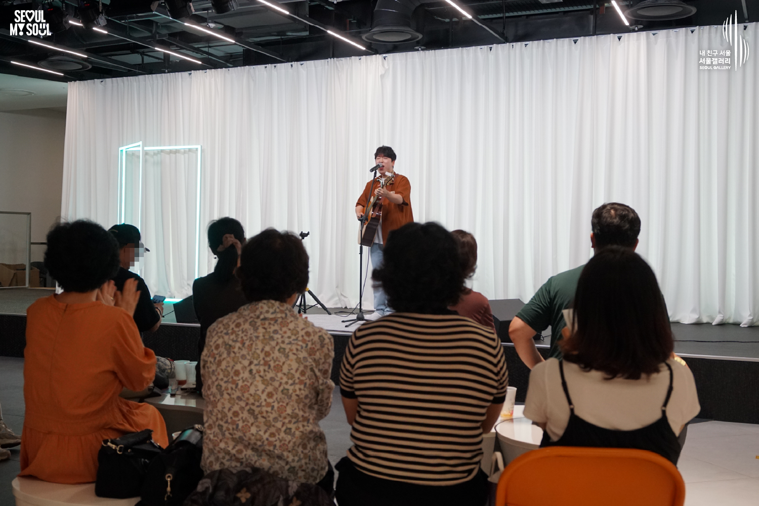 SEOUL MY SOUL 로고와 내 친구 서울 서울갤러리 SEOUL GALLERY 로고가 표기된 서울갤러리 공연장 사진으로, 천장 무대 조명과 푸른빛 사각 조명이 비추는 흰색 커튼 배경의 무대 위에서 짧은 머리에 셔츠와 바지를 입은 남성 싱어송라이터가 어쿠스틱 기타를 연주하며 마이크 앞에서 노래하고 있으며, 객석에는 여러 관람객들이 앉아 무대를 바라보는 모습이 담긴 이미지입니다.