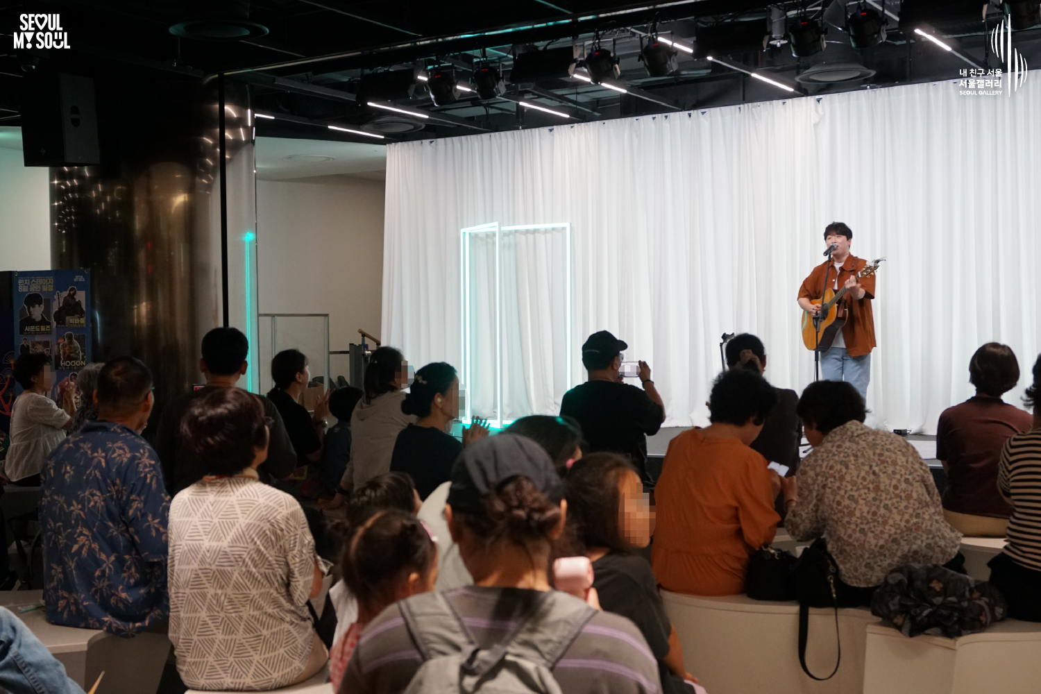 SEOUL MY SOUL 로고와 내 친구 서울 서울갤러리 SEOUL GALLERY 로고가 표기된 서울갤러리 공연장 사진으로, 천장 무대 조명과 푸른빛 사각 조명이 비추는 흰색 커튼 배경의 무대 위에서 짧은 머리에 셔츠와 바지를 입은 남성 싱어송라이터가 어쿠스틱 기타를 연주하며 노래하고 있으며, 객석에는 다양한 연령대의 관람객들이 앉아 공연을 관람하고 일부는 휴대전화로 무대를 촬영하는 모습이 담긴 이미지입니다.