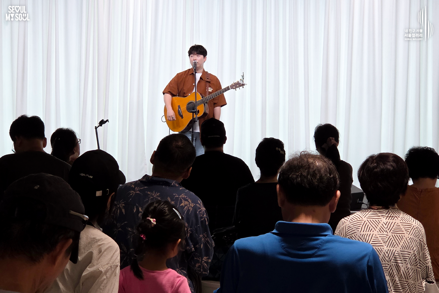 SEOUL MY SOUL 로고와 내 친구 서울 서울갤러리 SEOUL GALLERY 로고가 표기된 서울갤러리 공연장 사진으로, 천장 무대 조명이 비추는 흰색 커튼 배경의 무대 위에서 짧은 머리에 셔츠와 바지를 입은 남성 싱어송라이터가 어쿠스틱 기타를 메고 마이크 앞에서 노래하고 있으며, 객석 전경에는 어린이를 포함한 다양한 연령대의 관람객들이 앉아 무대를 바라보는 모습이 담긴 이미지입니다.