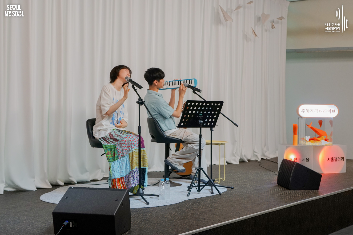 SEOUL MY SOUL 로고와 내 친구 서울 서울갤러리 SEOUL GALLERY 로고가 표기된 서울갤러리 공연장 무대 사진으로, 천장 조명이 비추고 종이비행기 장식이 달린 흰색 커튼 배경의 무대에서 짧은 머리의 여성 아티스트가 의자에 앉아 마이크 앞에서 노래하며 한 손을 들어 올리고 있고, 옆의 짧은 머리 남성 아티스트는 건반 하모니카를 들어 연주하고 있으며 앞에는 악보대와 모니터 스피커가 놓여 있는 모습이 담긴 이미지입니다.