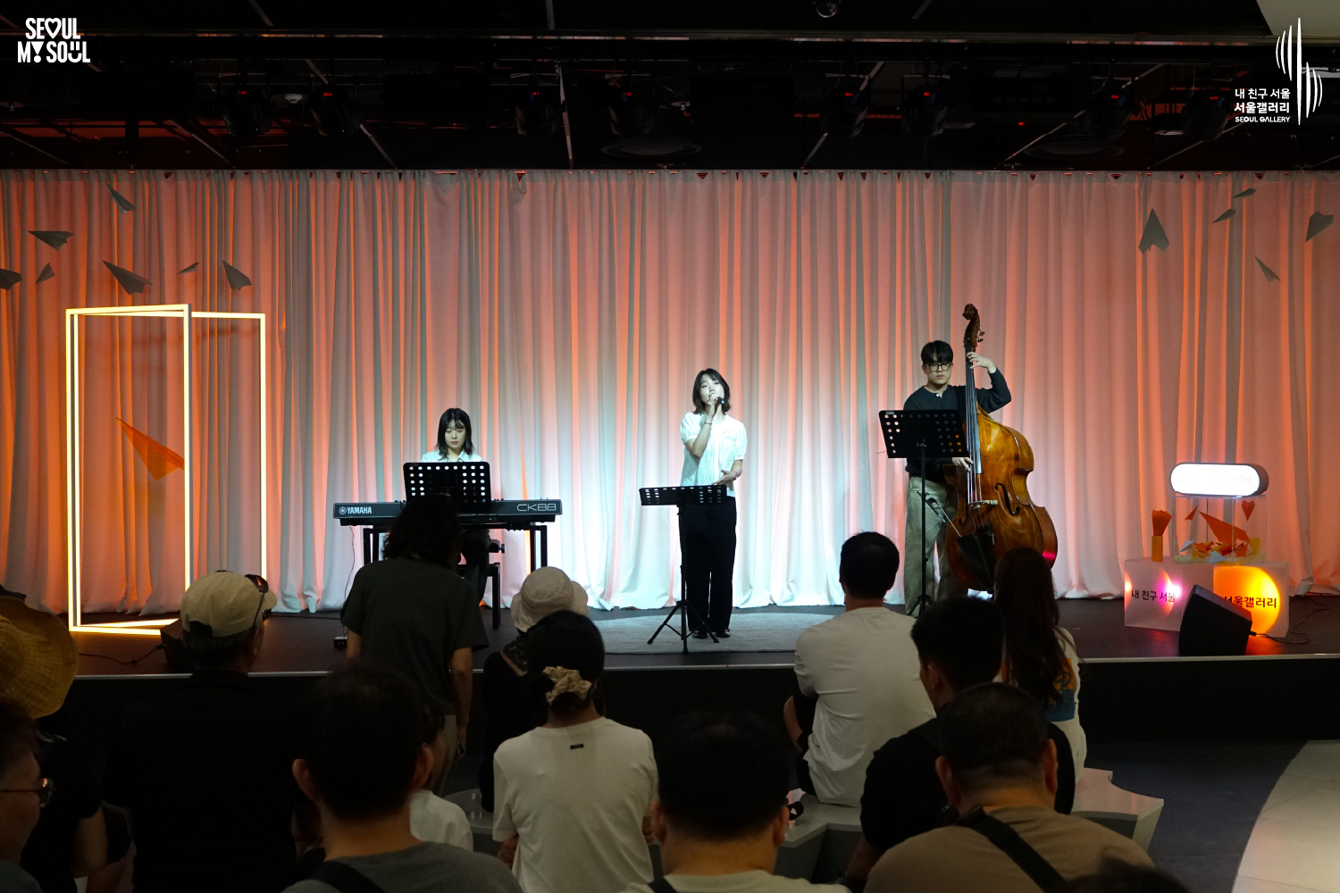 SEOUL MY SOUL 로고와 내 친구 서울 서울갤러리 SEOUL GALLERY 로고가 표기된 서울갤러리 공연장 사진으로, 주황빛과 붉은빛 조명이 비추는 흰색 커튼 배경의 무대에서 여성 보컬이 마이크를 들고 노래하고 있으며, 왼쪽의 여성 연주자는 키보드를 연주하고 오른쪽의 안경을 착용한 남성 연주자는 콘트라베이스를 연주하고 있고, 객석에는 여러 관람객들이 앉아 무대를 바라보는 모습이 담긴 이미지입니다.
