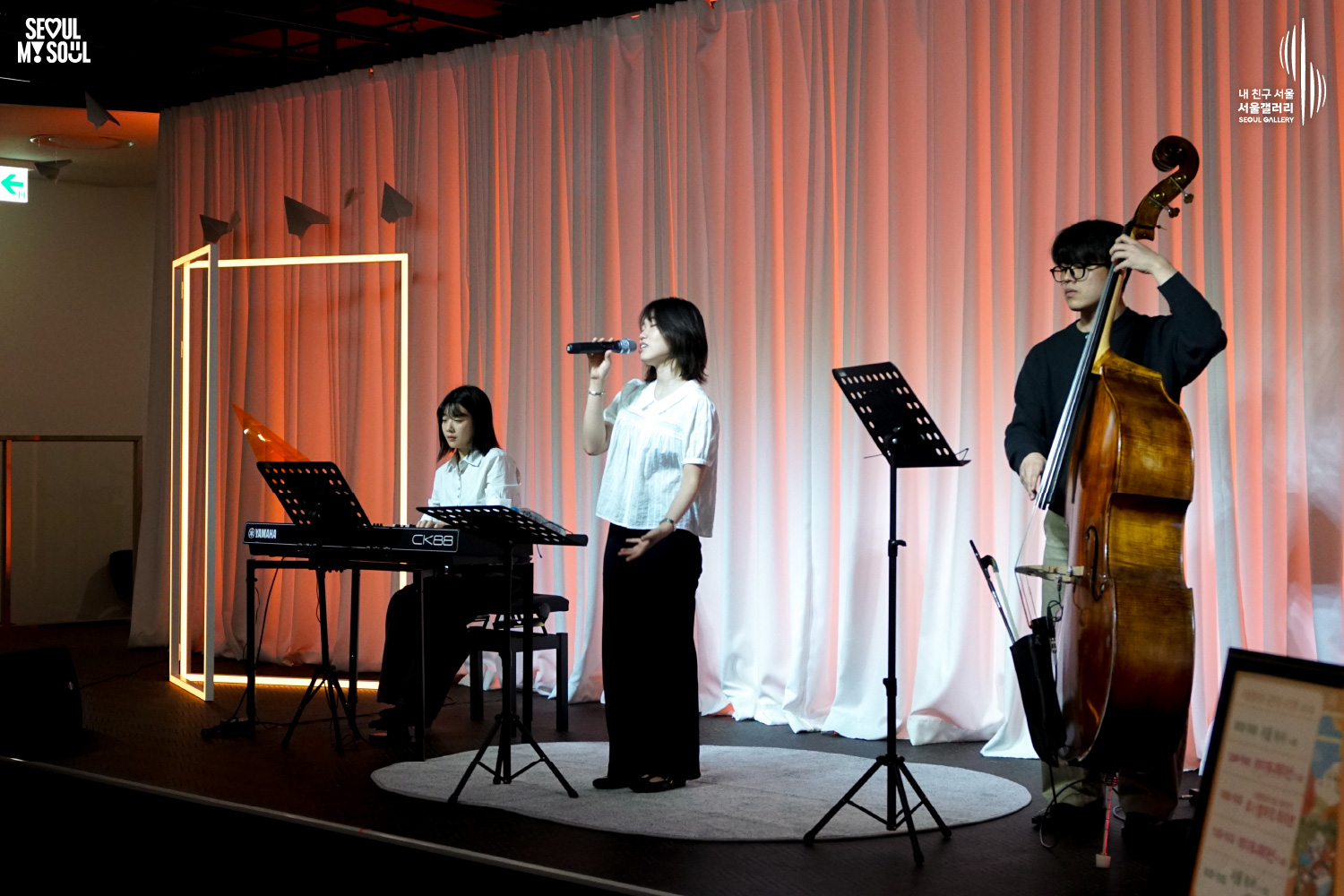 SEOUL MY SOUL 로고와 내 친구 서울 서울갤러리 SEOUL GALLERY 로고가 표기된 서울갤러리 공연장 무대 사진으로, 주황빛 조명이 비추고 종이비행기 장식과 사각 조명이 설치된 흰색 커튼 배경의 무대에서 단발머리 여성 보컬이 마이크를 들고 노래하고 있으며, 여성 연주자가 키보드를 연주하고 안경을 착용한 남성 연주자가 콘트라베이스를 연주하고 있으며, 앞에는 악보대가 놓여 있는 모습이 담긴 이미지입니다.