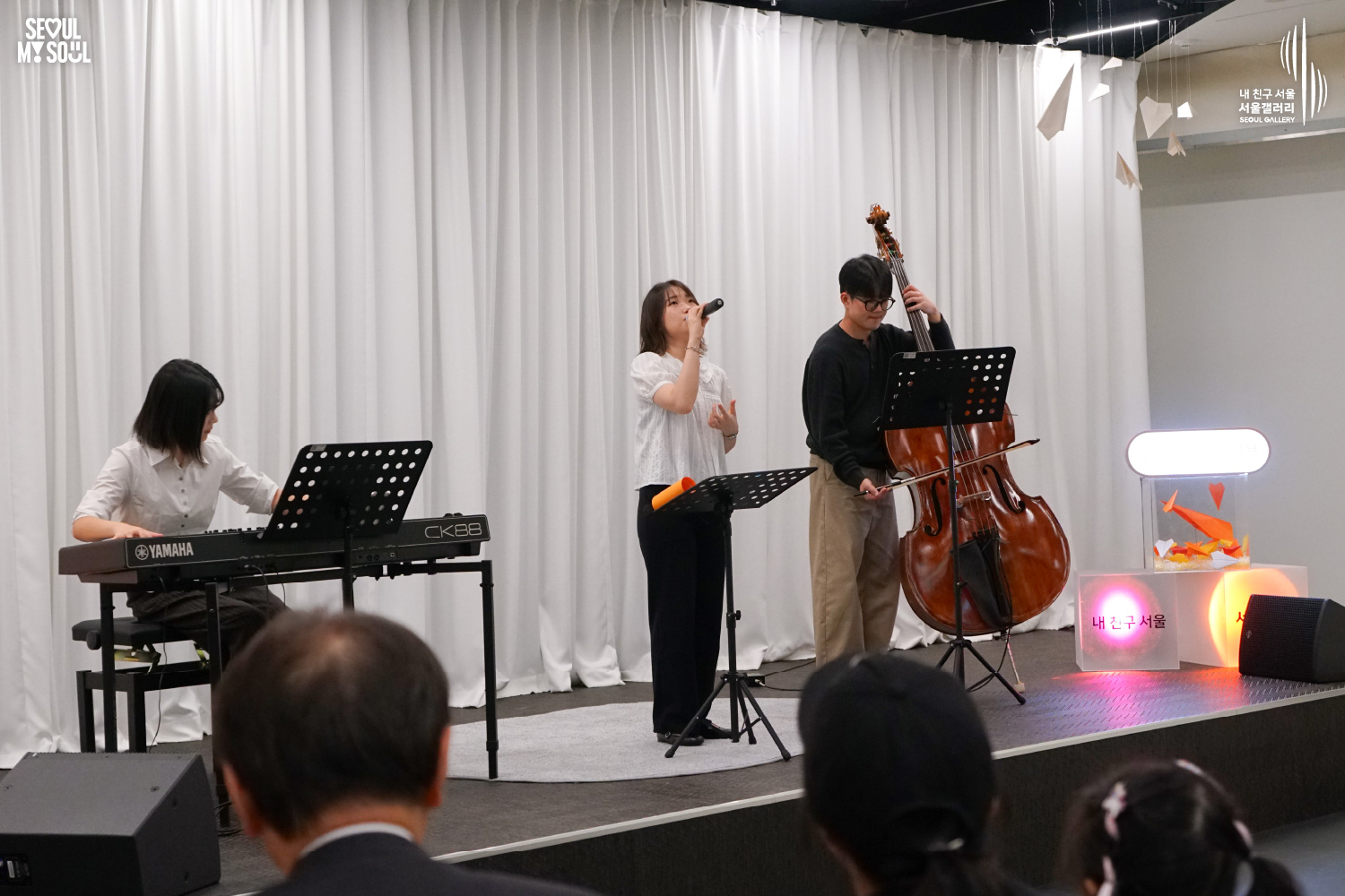 SEOUL MY SOUL 로고와 내 친구 서울 서울갤러리 SEOUL GALLERY 로고가 표기된 서울갤러리 공연장 사진으로, 천장 무대 조명이 비추는 흰색 커튼 배경의 무대에서 단발머리 여성 보컬이 마이크를 들고 노래하고 있으며, 왼쪽의 여성 연주자는 키보드를 연주하고 오른쪽의 안경을 착용한 남성 연주자는 콘트라베이스를 연주하고 있고, 무대에는 악보대와 모니터 스피커가 놓여 있으며 전경에는 관람객들의 뒷모습이 함께 담긴 이미지입니다.