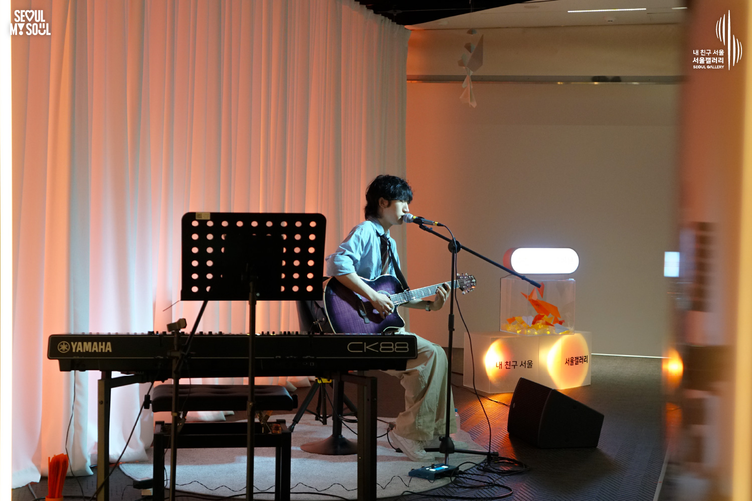 SEOUL MY SOUL 로고와 내 친구 서울 서울갤러리 SEOUL GALLERY 로고가 표기된 서울갤러리 공연장 무대 사진으로, 주황빛 조명이 비추는 흰색 커튼 배경의 무대에서 웨이브 머리에 셔츠와 바지를 입은 남성 싱어송라이터가 의자에 앉아 어쿠스틱 기타를 연주하며 마이크 앞에서 노래하고 있으며, 주변에는 키보드와 악보대, 모니터 스피커가 놓여 있는 모습이 담긴 이미지입니다.