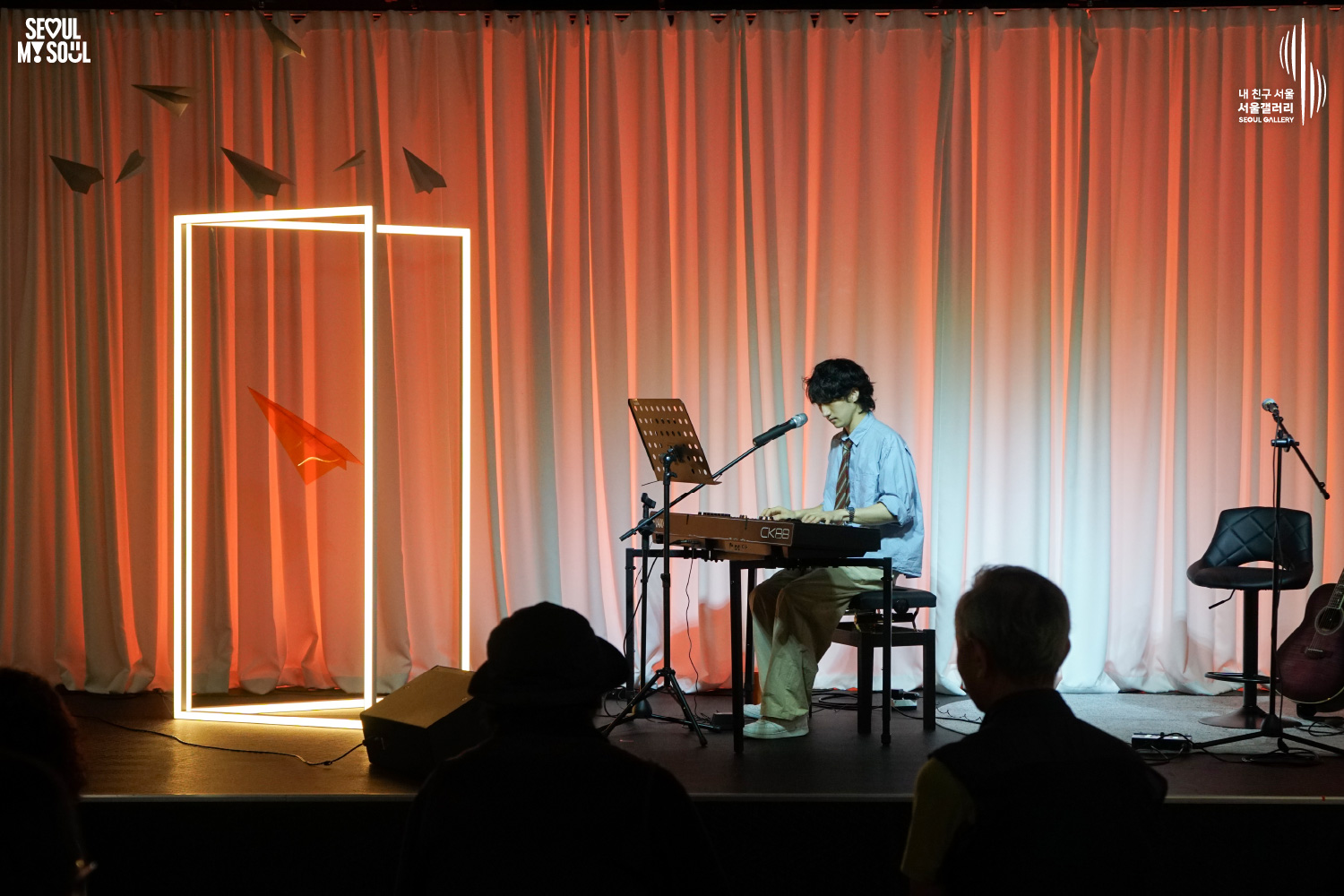 SEOUL MY SOUL 로고와 내 친구 서울 서울갤러리 SEOUL GALLERY 로고가 표기된 서울갤러리 공연장 사진으로, 주황빛과 붉은빛 조명과 사각 조명이 비추는 흰색 커튼 배경의 무대에서 웨이브 머리에 셔츠와 바지를 입은 남성 아티스트가 의자에 앉아 키보드를 양손으로 연주하고 있으며, 앞에는 마이크와 악보대가 놓여 있고 오른쪽에는 기타와 의자, 마이크 스탠드가 있으며 전경에는 관람객들의 뒷모습이 담긴 이미지입니다.