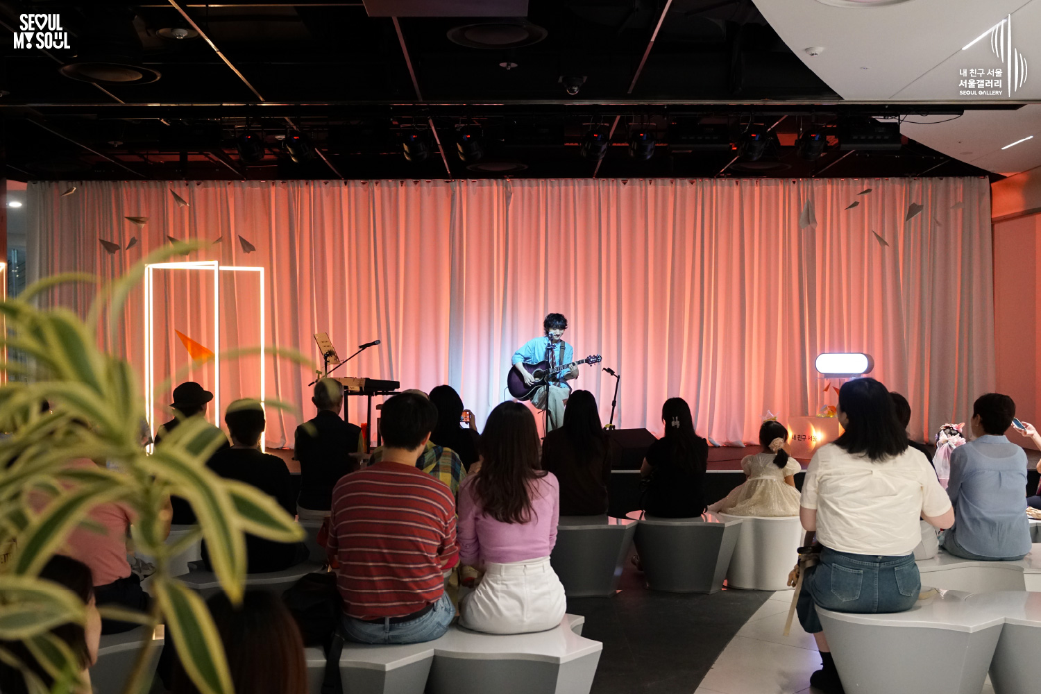 SEOUL MY SOUL 로고와 내 친구 서울 서울갤러리 SEOUL GALLERY 로고가 표기된 서울갤러리 지하 1층 공연장의 공연 현장 사진으로, 주황빛 무대 조명이 비추는 흰색 커튼 배경의 무대에서 웨이브 머리에 셔츠와 바지를 입은 남성 싱어송라이터가 의자에 앉아 어쿠스틱 기타를 연주하며 노래하고 있으며, 객석에는 어린이를 포함한 다양한 연령대의 관람객들이 앉아 무대를 바라보는 모습이 담긴 이미지입니다.