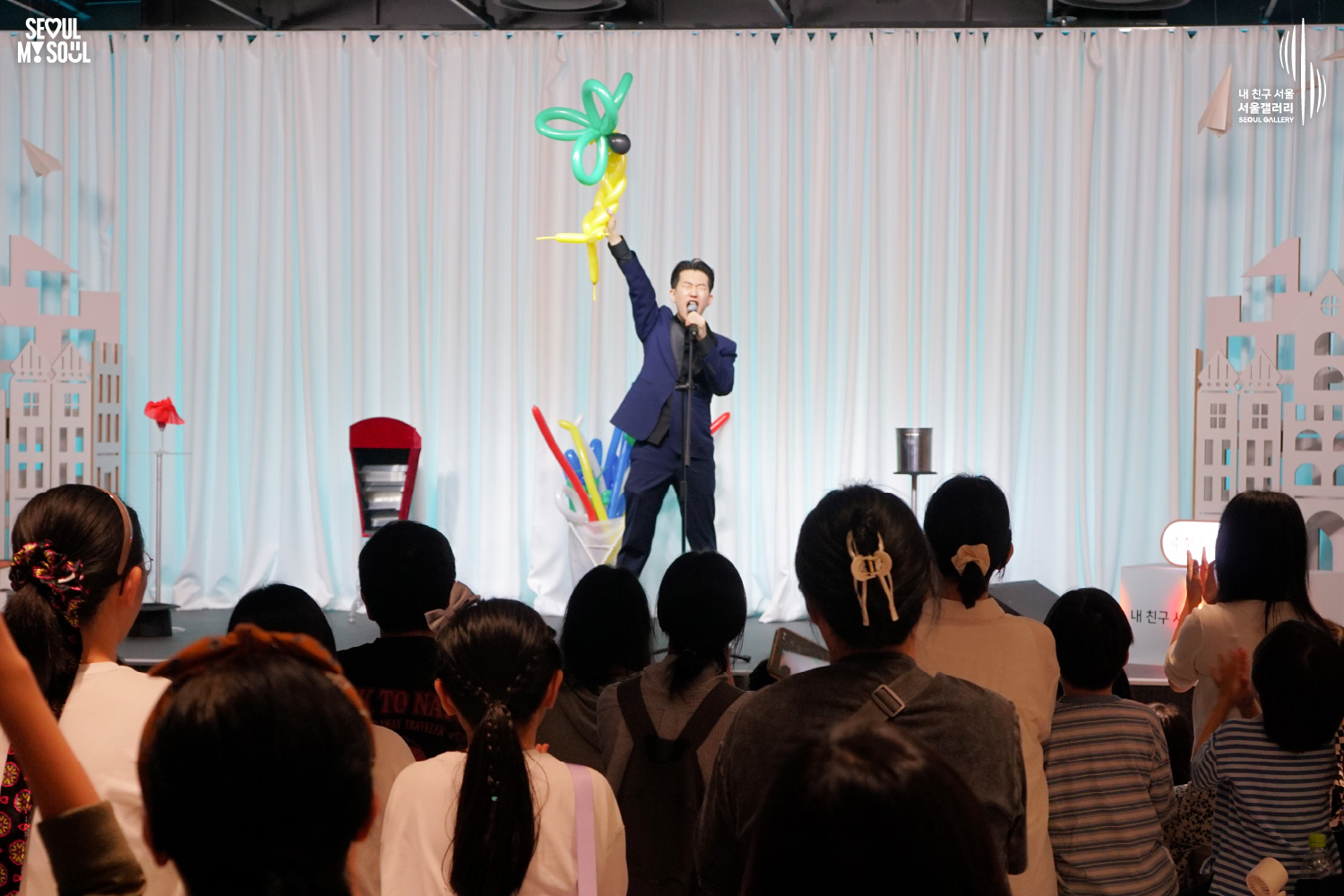 SEOUL MY SOUL 로고와 내 친구 서울 서울갤러리 SEOUL GALLERY 로고가 표기된 서울갤러리 공연장 사진으로, 푸른빛 조명이 비추는 흰색 커튼 배경의 무대 중앙에서 정장을 입은 남성 마술사 도재혁이 마이크를 들고 커다란 풍선 조형물을 머리 위로 높이 들어 올리고 있으며, 객석 전경에는 어린아이와 성인 관람객들이 무대를 바라보거나 박수를 치는 모습이 담긴 이미지입니다.