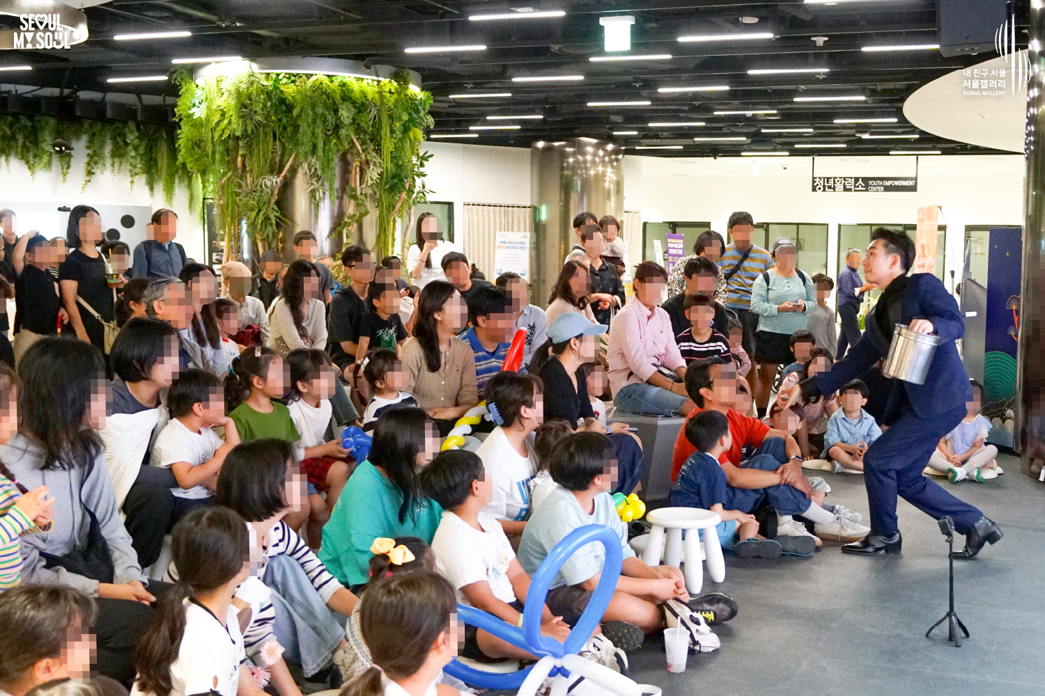 SEOUL MY SOUL 로고와 내 친구 서울 서울갤러리 SEOUL GALLERY 로고가 표기된 서울갤러리 지하 1층 공연장의 공연 현장 사진으로, 천장 조명이 밝게 비추는 객석 앞에서 정장을 입은 남성 마술사 도재혁이 금속 원통형 소품을 한 손에 들고 관람객 가까이에서 공연을 펼치고 있으며, 어린아이들과 성인 관람객들이 둘러앉아 무대를 바라보고 풍선 조형물을 들고 있는 모습이 담긴 이미지입니다.