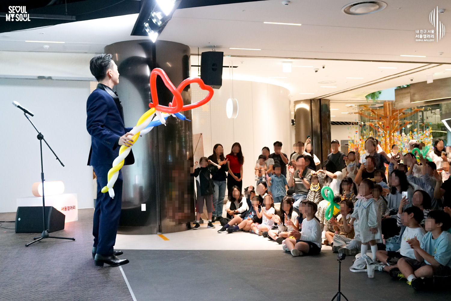 SEOUL MY SOUL 로고와 내 친구 서울 서울갤러리 SEOUL GALLERY 로고가 표기된 서울갤러리 공연장 사진으로, 천장 조명이 밝게 비추는 실내 공간에서 정장을 입은 남성 마술사 도재혁이 하트 모양 풍선이 연결된 긴 풍선 조형물을 들고 관객을 바라보고 있으며, 무대 앞에는 어린아이를 포함한 관람객들이 앉거나 서서 박수를 치고 손을 들며 공연을 관람하는 모습이 담긴 이미지입니다.