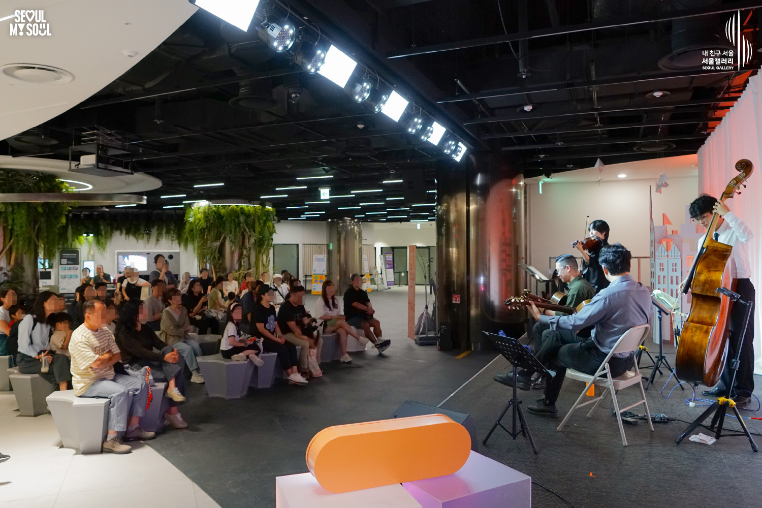 SEOUL MY SOUL 로고와 내 친구 서울 서울갤러리 SEOUL GALLERY 로고가 표기된 서울갤러리 지하 1층 공연장의 공연 현장 사진으로, 천장 무대 조명과 주황빛 조명이 비추는 무대에서 라쁘띠프랑스콰르텟의 네 연주자가 바이올린과 더블베이스, 기타를 연주하고 있으며, 무대 옆 넓은 객석에는 다양한 연령대의 관람객들이 앉아 공연을 관람하는 모습이 담긴 이미지입니다.