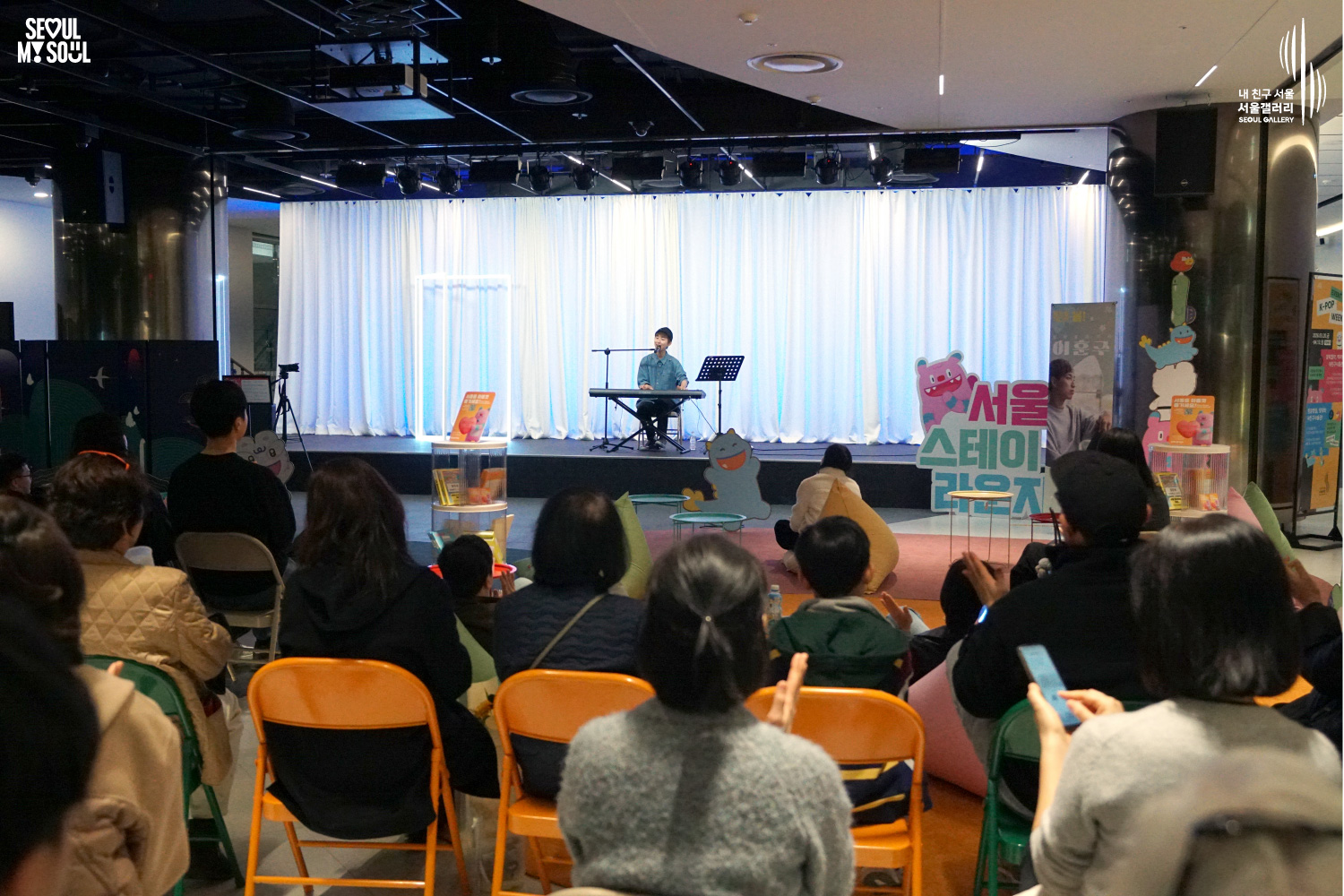 Solo musician Lee Hojun performs vocals and keyboard at Seoul Gallery's Seoul Stage Lounge event before a large seated audience, with "SEOUL MY SOUL" and "Seoul Gallery" logos visible.