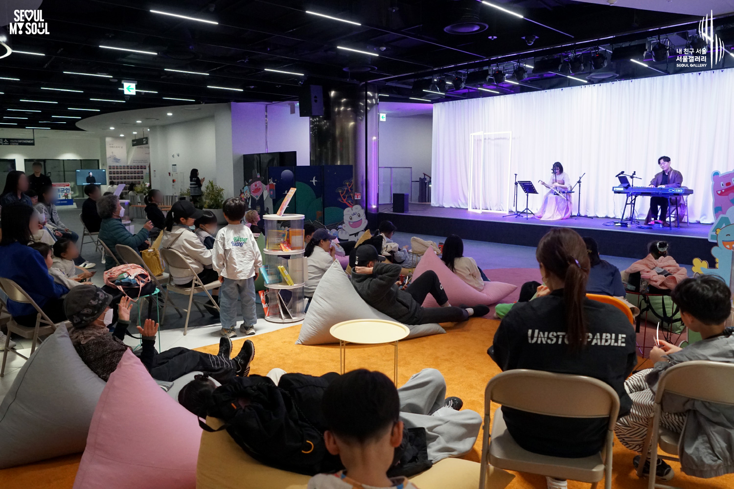 A haegeum and keyboard duo performs at Seoul Gallery before a large audience of all ages relaxing on chairs and beanbags, with "SEOUL MY SOUL" and "Seoul Gallery" logos visible.