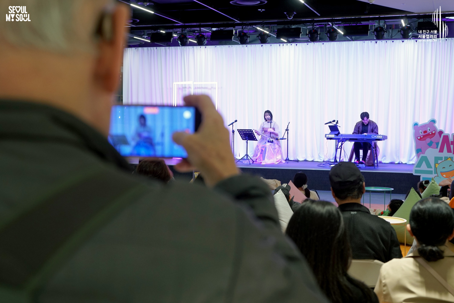 An audience member films a haegeum and keyboard duo's performance at Seoul Gallery's Seoul Stage Lounge event, with "SEOUL MY SOUL" and "Seoul Gallery" logos visible.