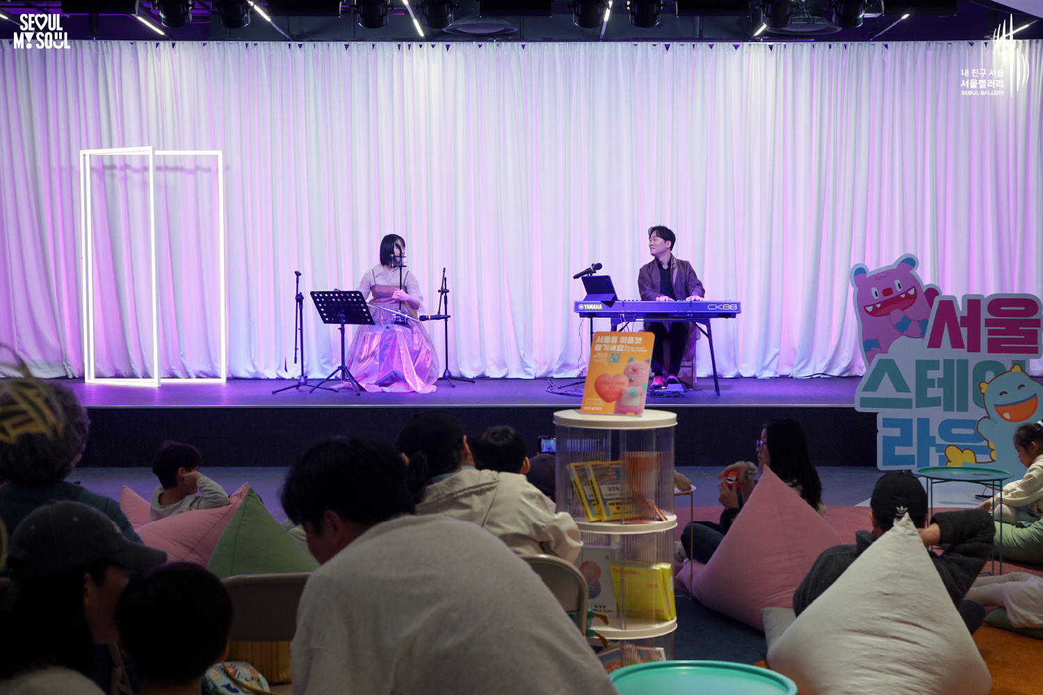 A haegeum player in a pink hanbok and a Yamaha keyboard player perform at Seoul Gallery's Seoul Stage Lounge event while audience members relax on beanbag chairs, with "SEOUL MY SOUL" and "Seoul Gallery" logos visible.