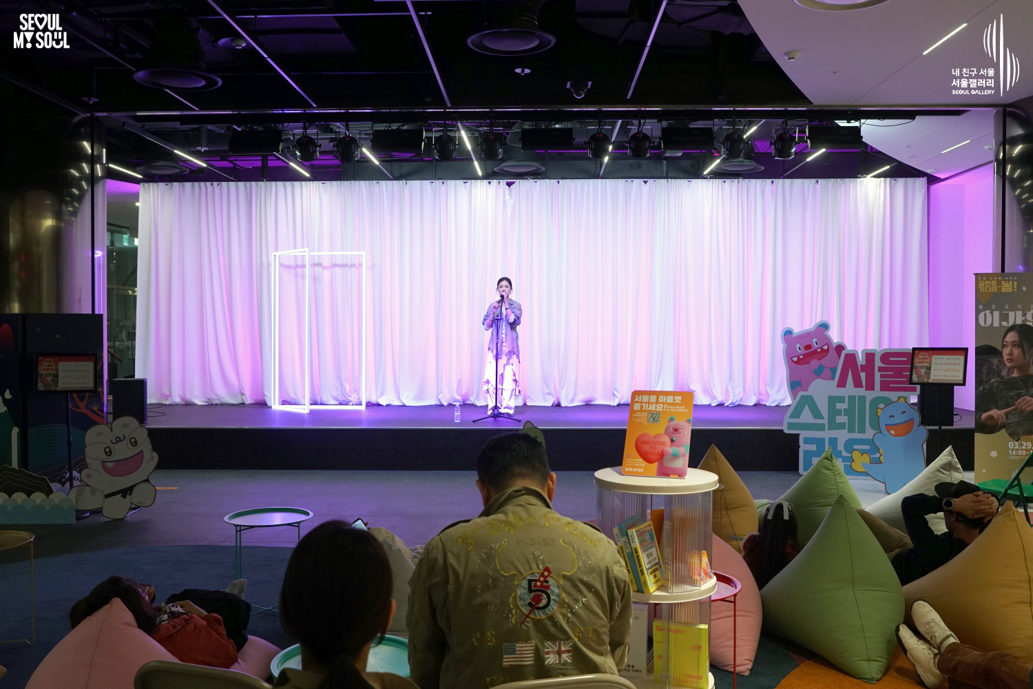 Vocalist Lee Gahyeon performs on a pink-lit stage at Seoul Gallery's Seoul Stage Lounge event while audience members relax on beanbag chairs, with "SEOUL MY SOUL" and "Seoul Gallery" logos visible.