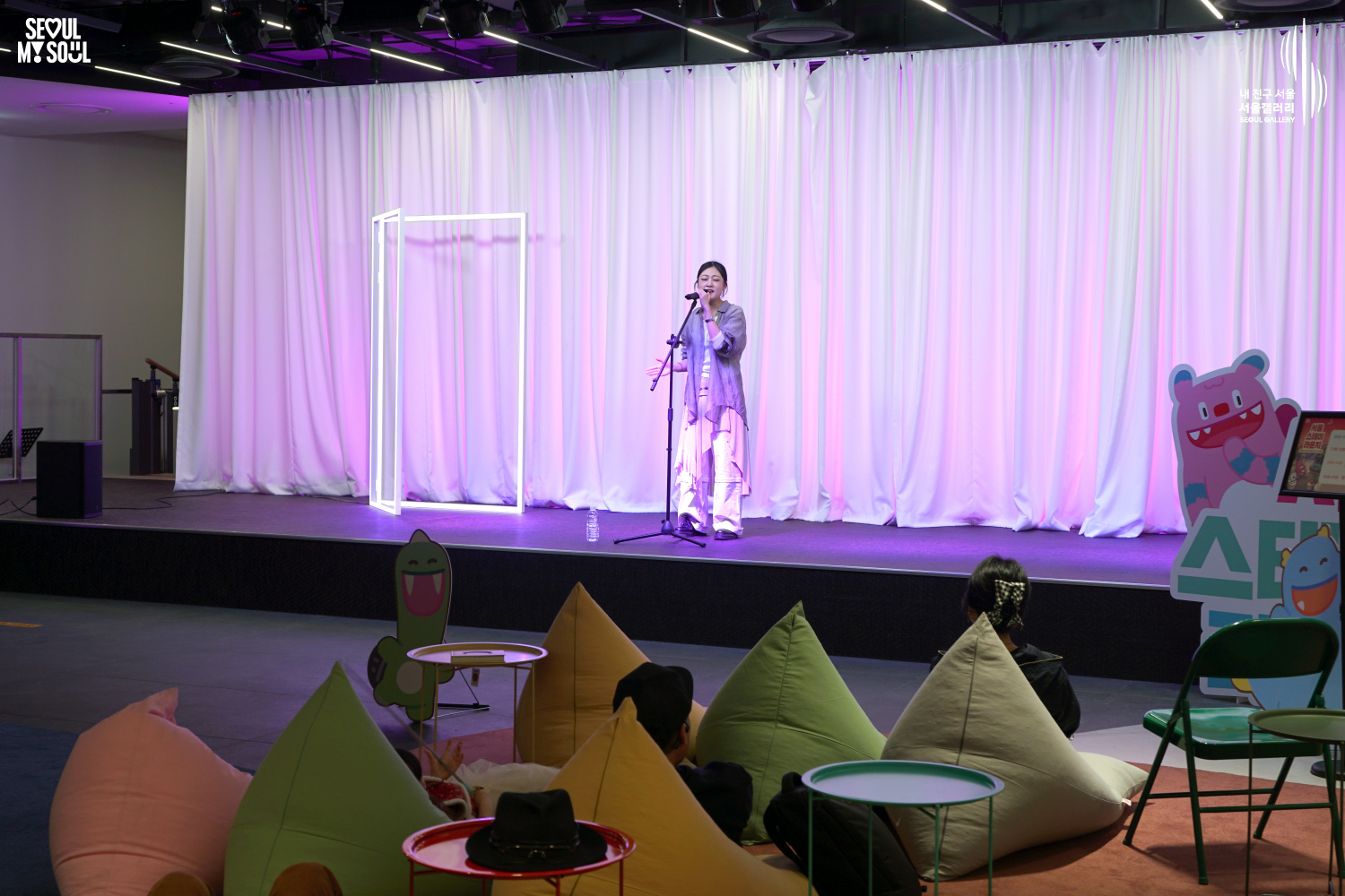 A female vocalist performs on a pink-lit stage at Seoul Gallery's Seoul Stage Lounge event while audience members relax on beanbag chairs, with "SEOUL MY SOUL" and "Seoul Gallery" logos visible.