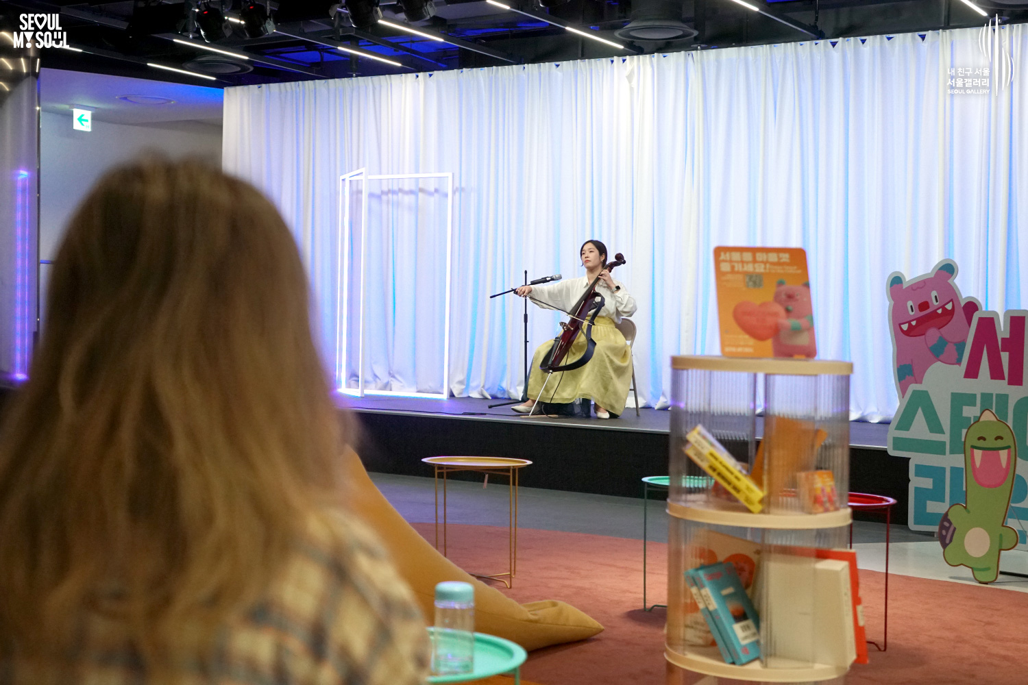 A female cellist in a yellow hanbok-style skirt performs at Seoul Gallery's Seoul Stage Lounge event, with "SEOUL MY SOUL" and "Seoul Gallery" logos visible.