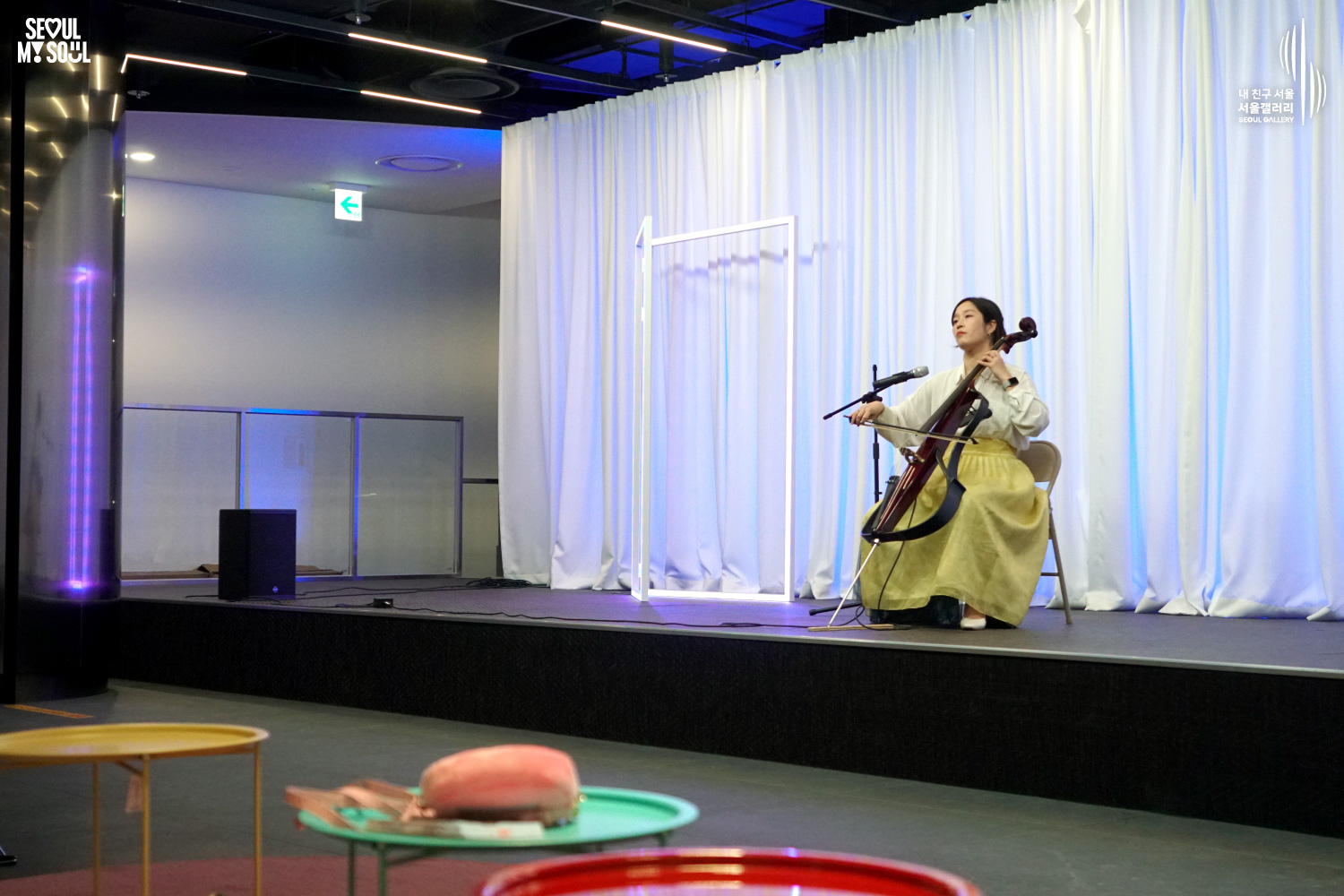 A female cellist in a yellow hanbok-style skirt performs on a blue-lit stage with white curtains at Seoul Gallery, with "SEOUL MY SOUL" and "Seoul Gallery" logos visible.