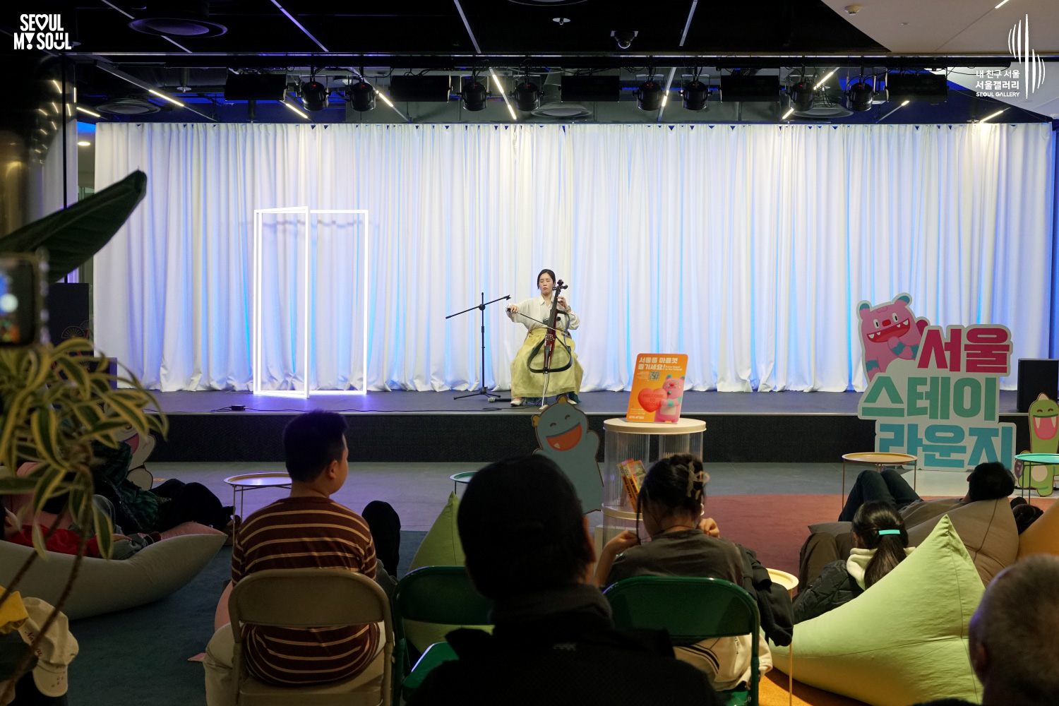Cellist Haru performs at Seoul Gallery's Seoul Stage Lounge event while audience members watch from beanbags and chairs, with "SEOUL MY SOUL" and "Seoul Gallery" logos visible.