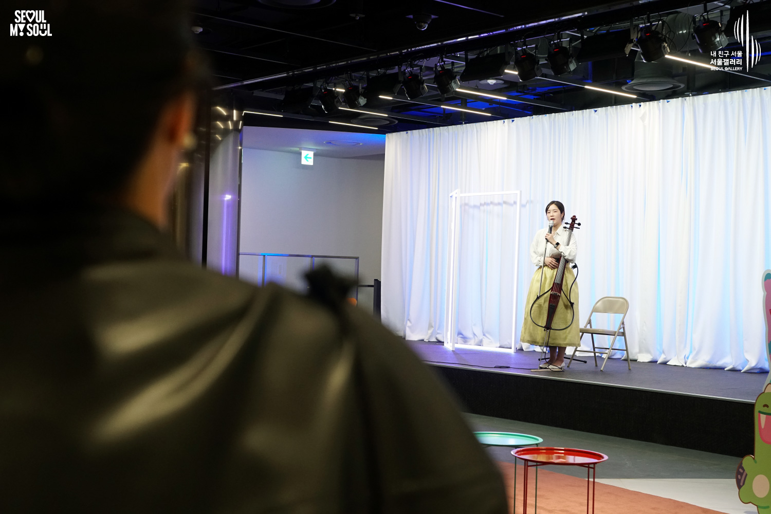 A female cellist in a yellow hanbok-style skirt speaks to the audience while standing with her cello on stage at Seoul Gallery, with "SEOUL MY SOUL" and "Seoul Gallery" logos visible.