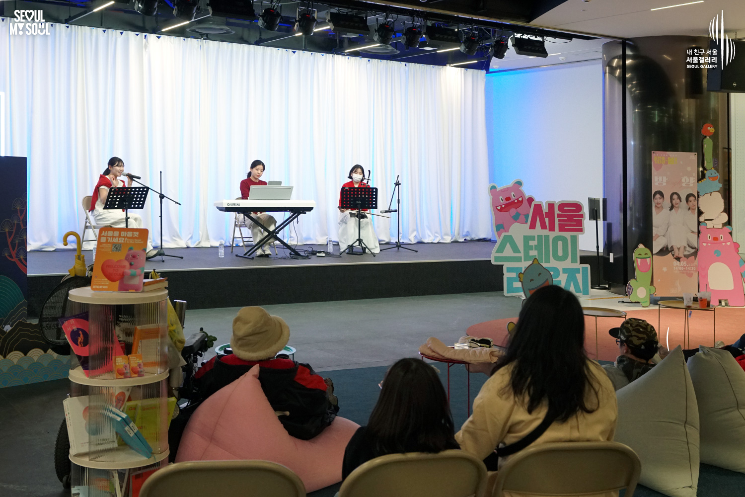 A female trio in red and white performs vocals and keyboard at Seoul Gallery's Seoul Stage event before an audience on beanbag chairs, with "SEOUL MY SOUL" and "Seoul Gallery" logos visible.