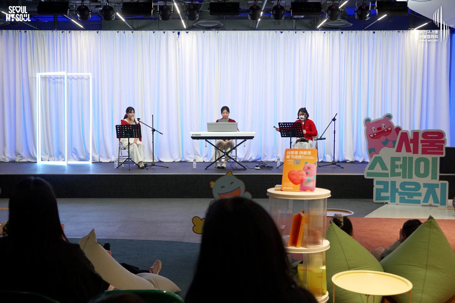 A female trio performs vocals and keyboard at Seoul Gallery's Seoul Stage Lounge event while audience members relax on beanbag chairs, with "SEOUL MY SOUL" and "Seoul Gallery" logos visible.