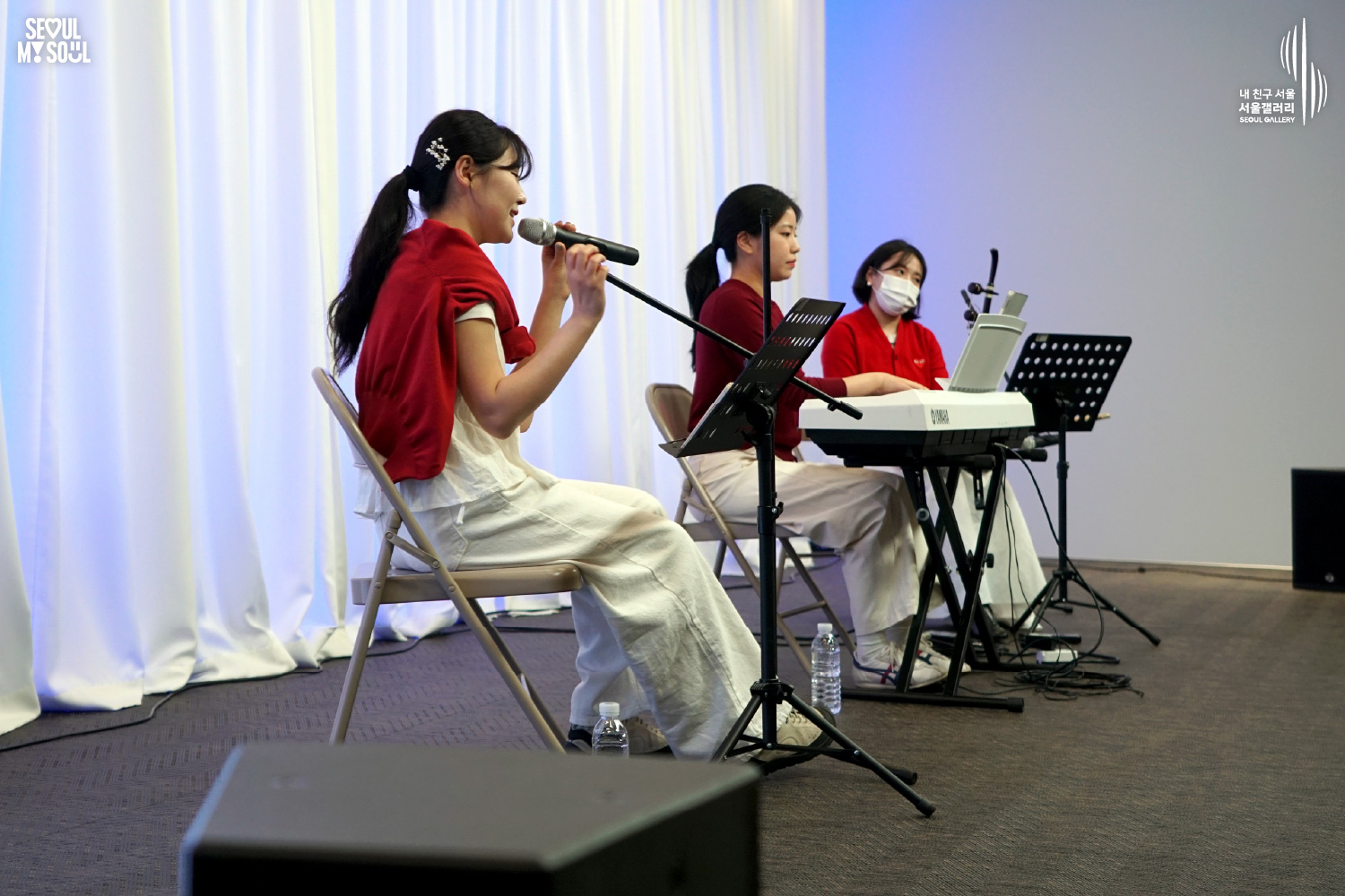 A female trio of two vocalists and a Yamaha keyboard player performs on a white-curtained stage at Seoul Gallery, with "SEOUL MY SOUL" and "Seoul Gallery" logos visible.